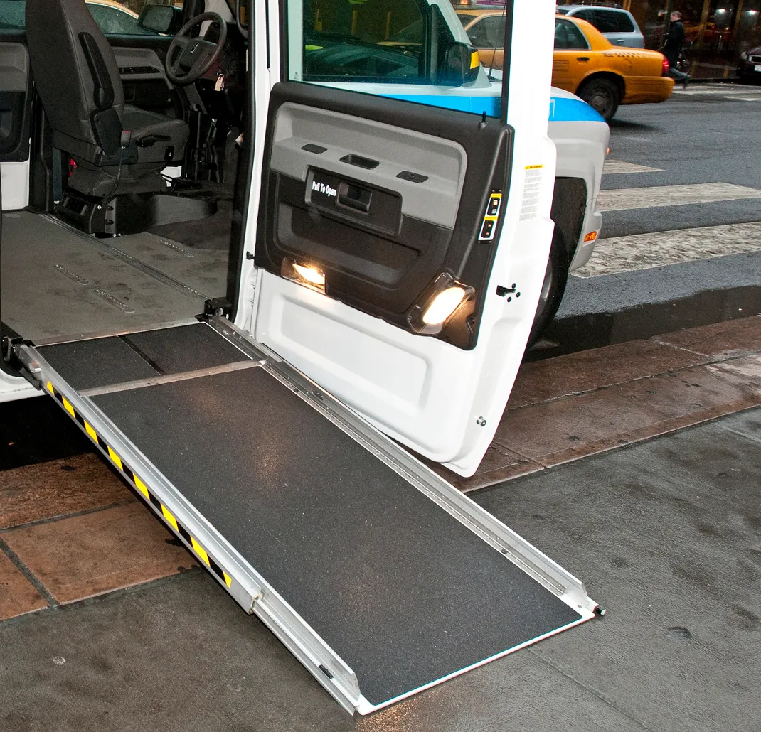 The open side door of a white wheelchair accessible van, with a fold-out ramp lowered from the van floor to a city sidewalk