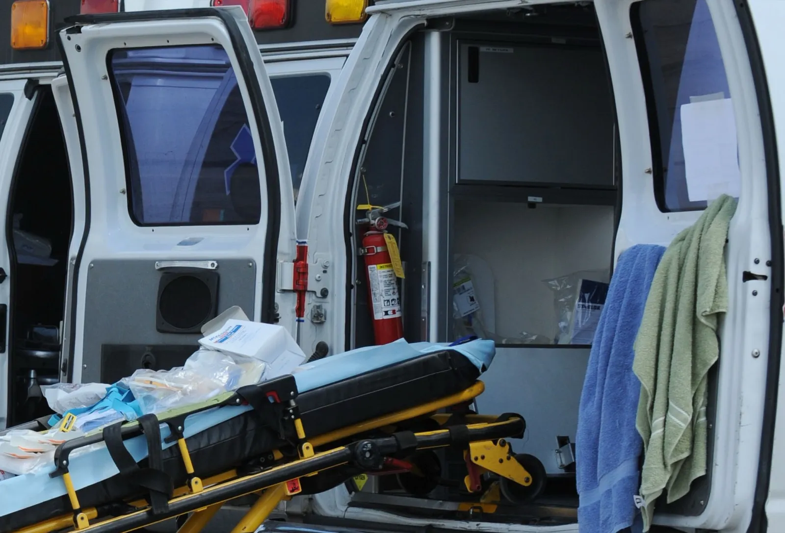 A wheeled ambulance stretcher with supplies on it stands beside the open rear doors of a white ambulance