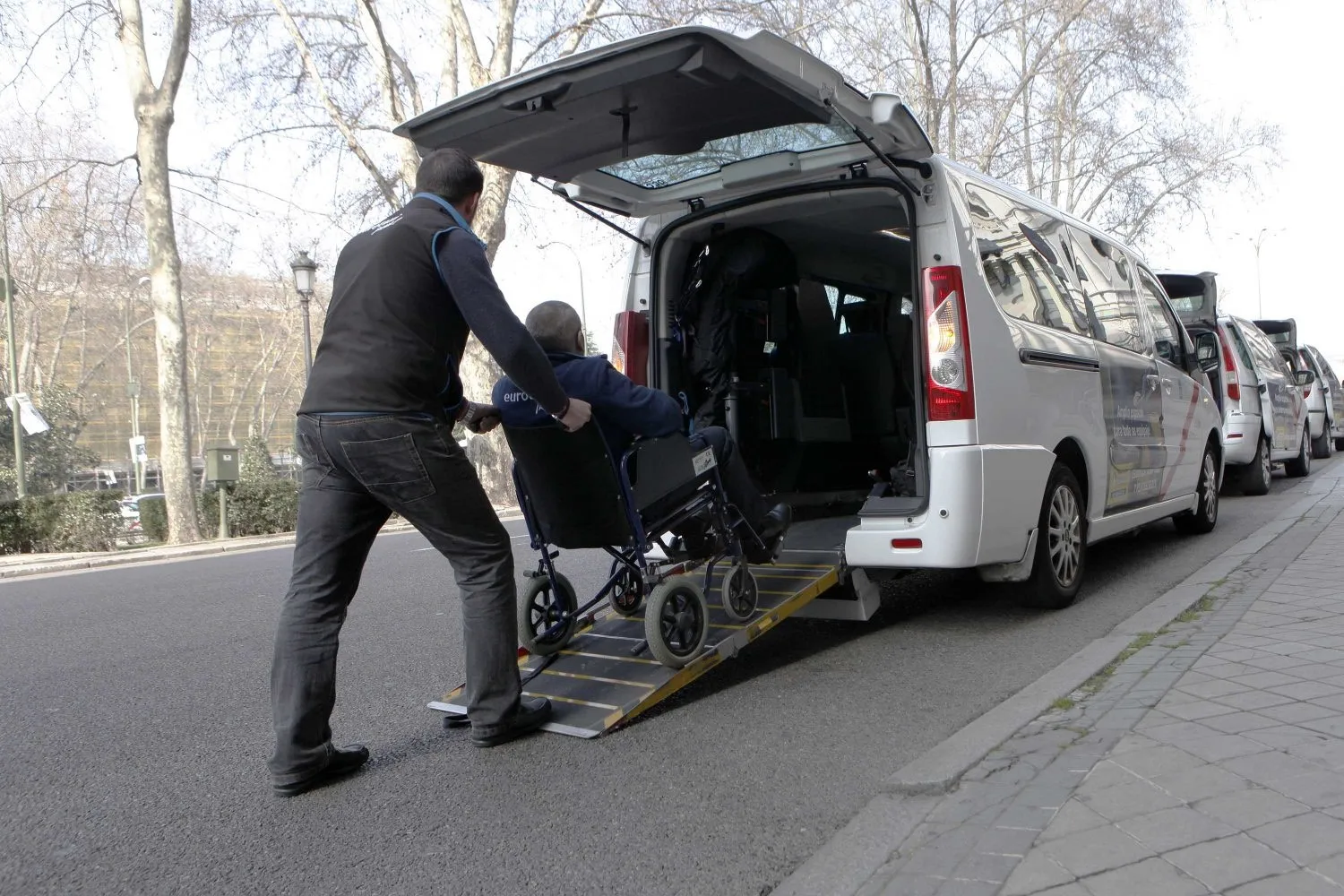 A taxi driver pushing a rider in a wheelchair up the rear ramp of a white accessible taxi van at the curb