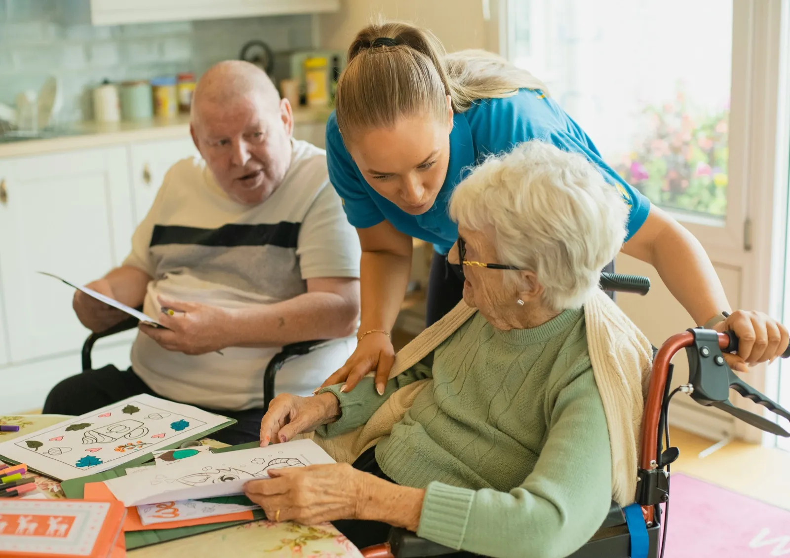 A caregiver leans over to help an older woman in a wheelchair with a coloring page at a table, while an older man holds his own page beside her