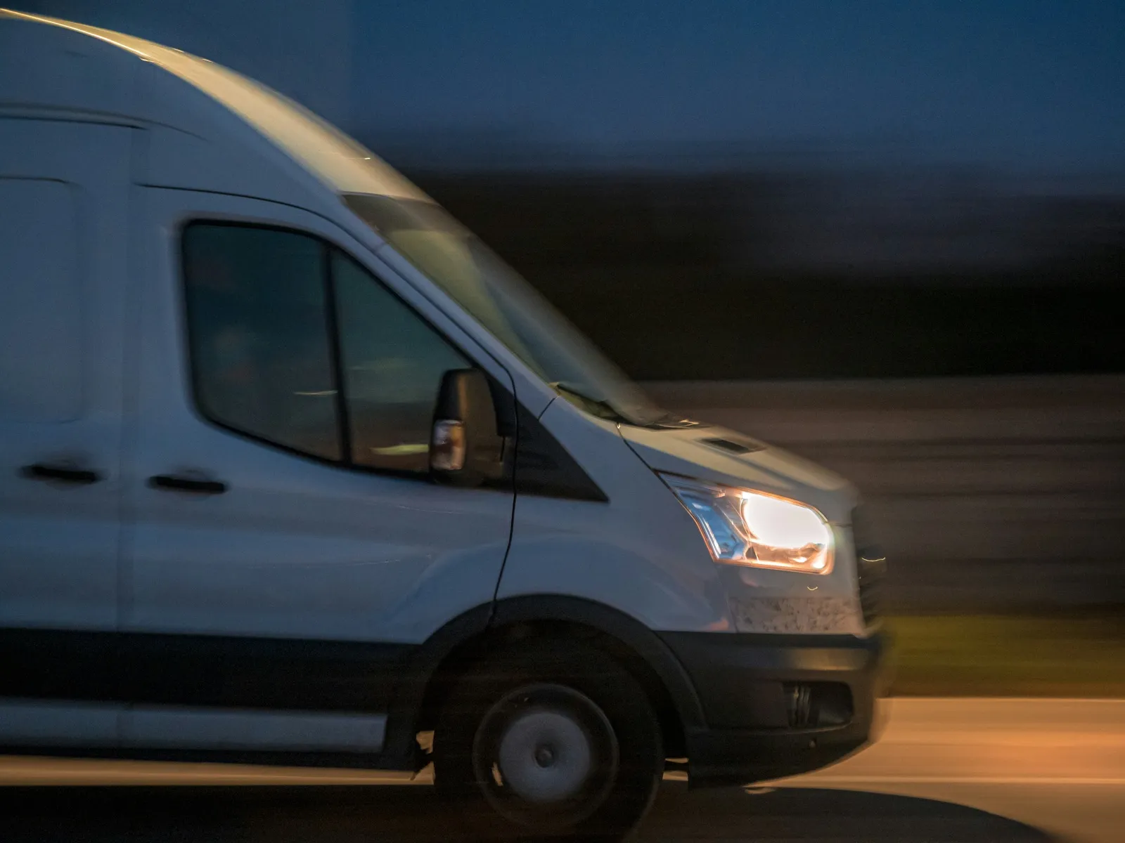 A white high-roof van driving with its headlights on at dusk