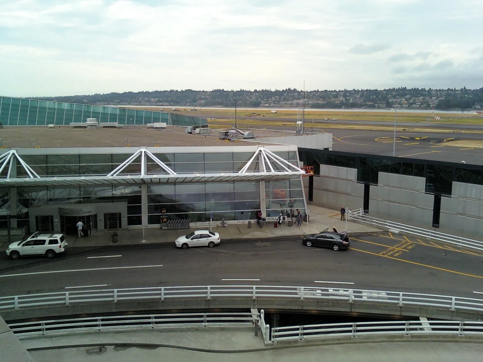 Cars stopped at the curb of a covered airport pickup and drop-off area, with travelers and luggage on the sidewalk and a runway behind the terminal
