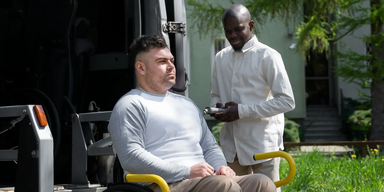A man in a wheelchair sits on the wheelchair lift of a van while an attendant stands beside him at the open door