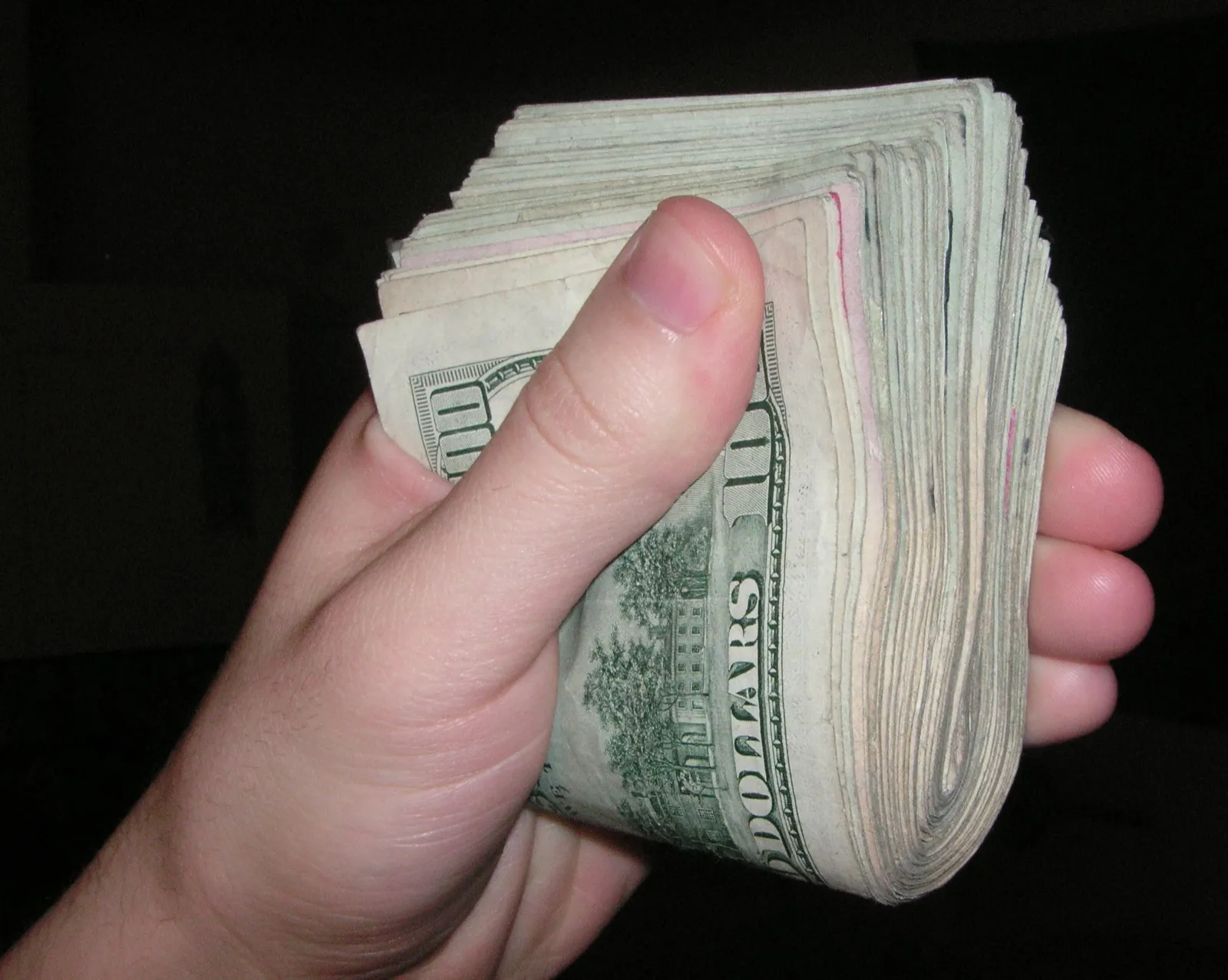 A hand gripping a thick fold of U.S. dollar bills, with hundred-dollar bills on the outside, against a black background