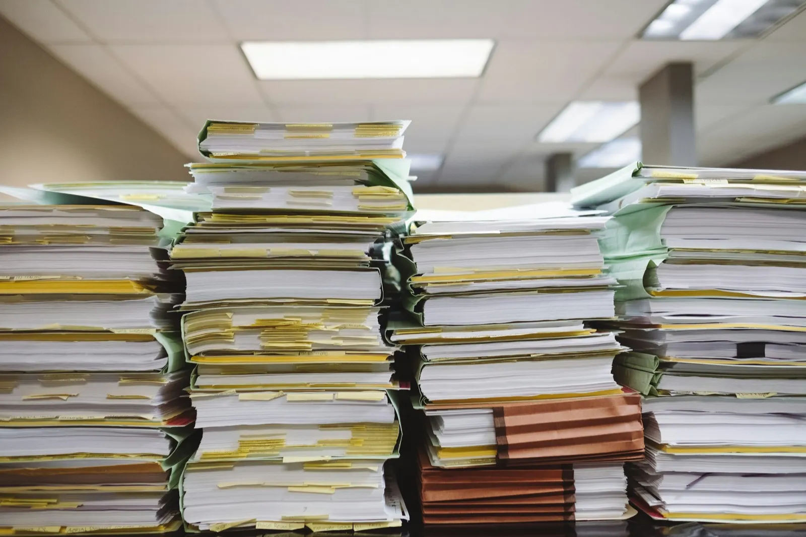Tall stacks of paper files with yellow and green tabs on a desk in an office