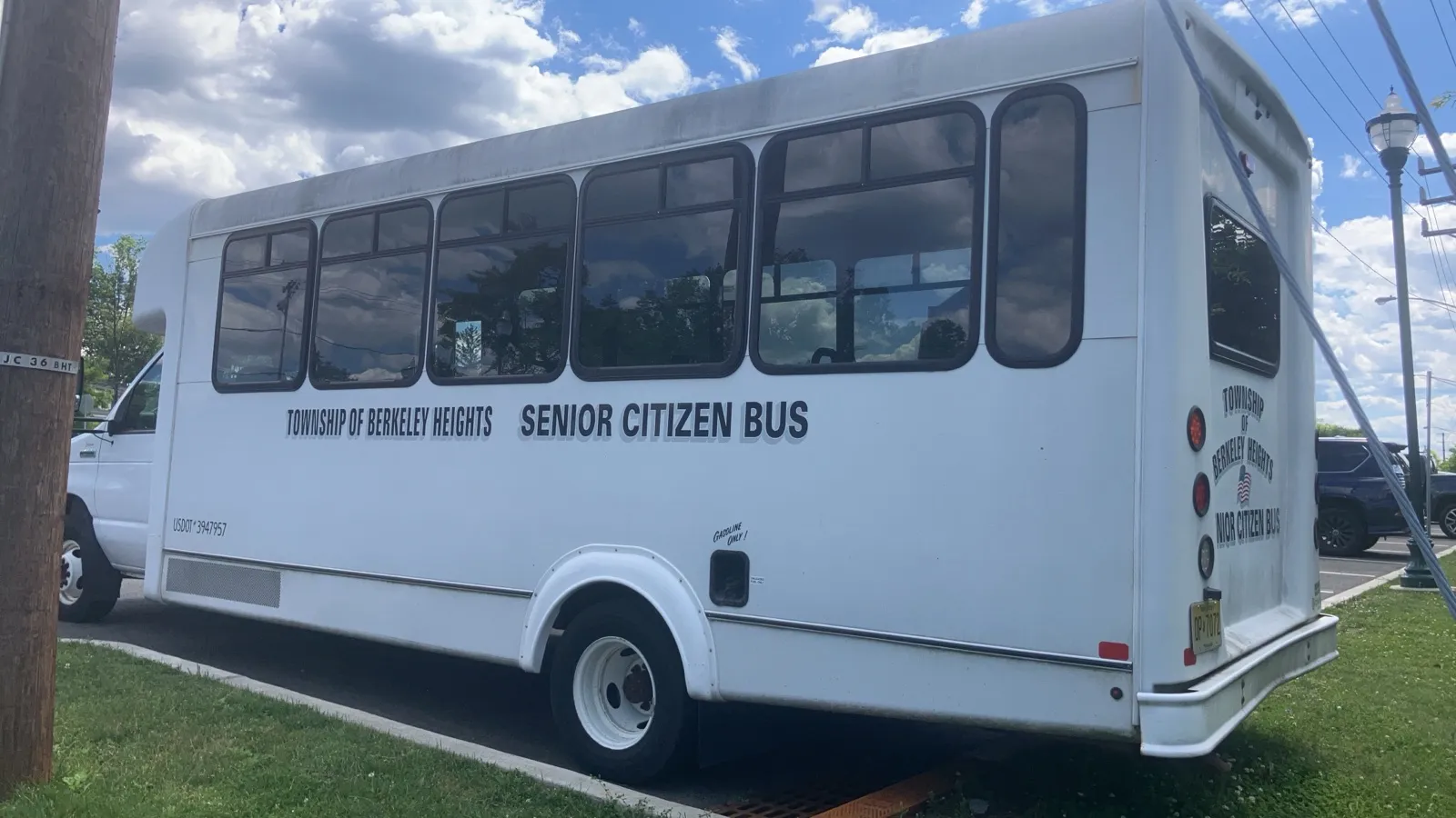 A white township senior citizen bus with a long row of side windows parked beside a grassy curb on a sunny day