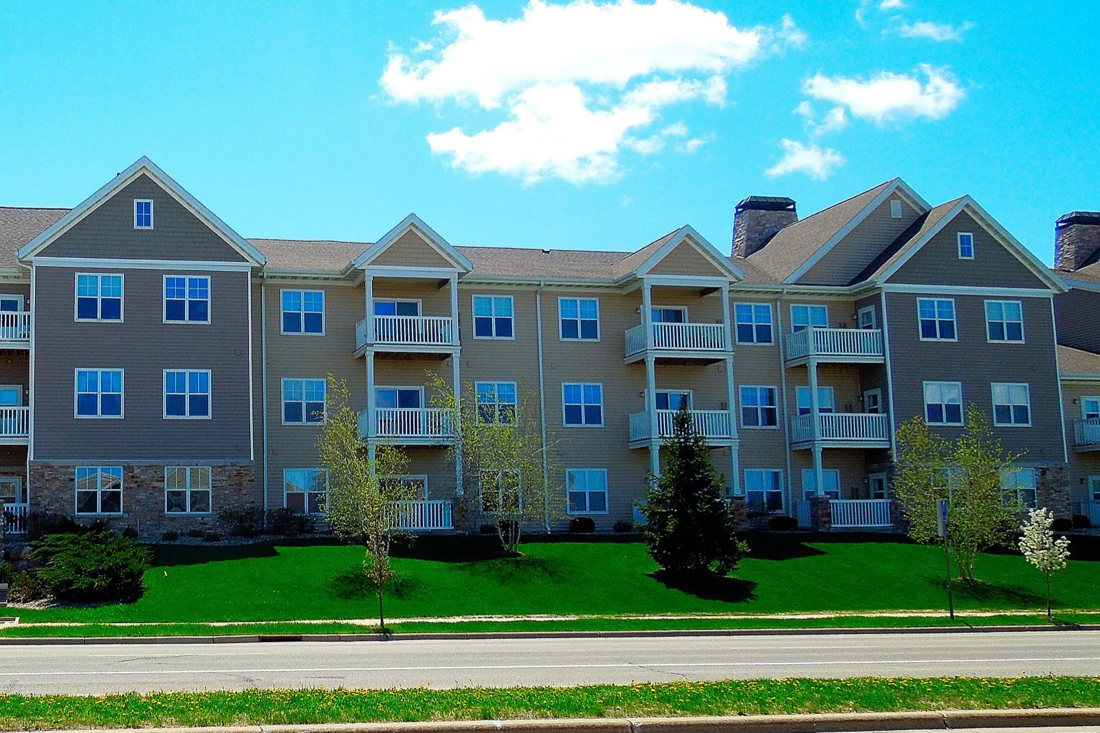 A three-story assisted living building with balconies and a sloping green lawn beside a quiet street, under a blue sky