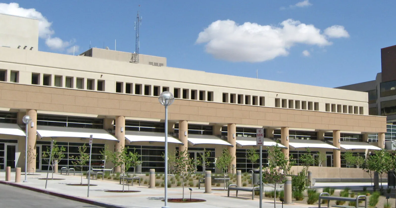 A hospital building in Albuquerque with a covered walkway, benches, and young trees under a blue sky