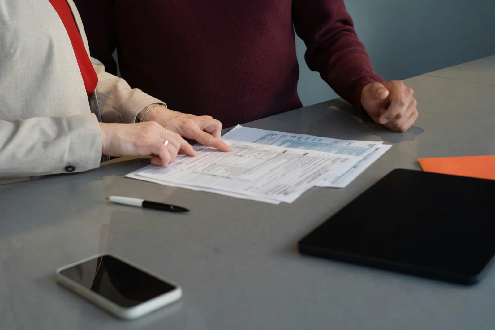 Two people at a table as one traces a line on a printed form with a finger, beside a pen, a phone, and a tablet