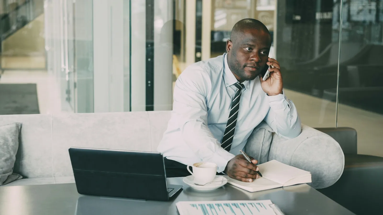 A man in a shirt and tie talks on his phone while writing notes in a notebook, with a laptop and a coffee cup on the table