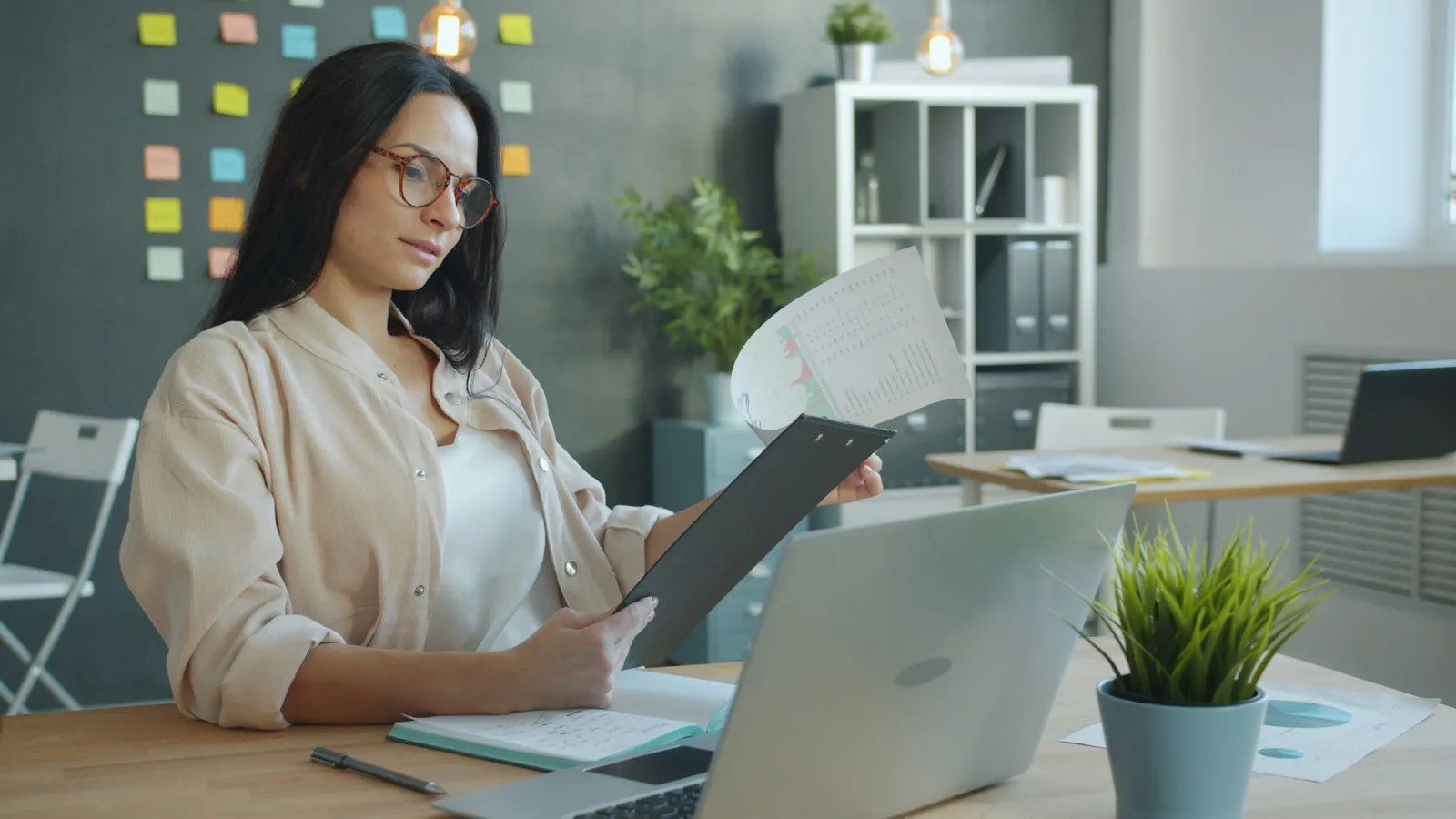 A woman at an office desk reads a printed sheet on a clipboard next to her open laptop