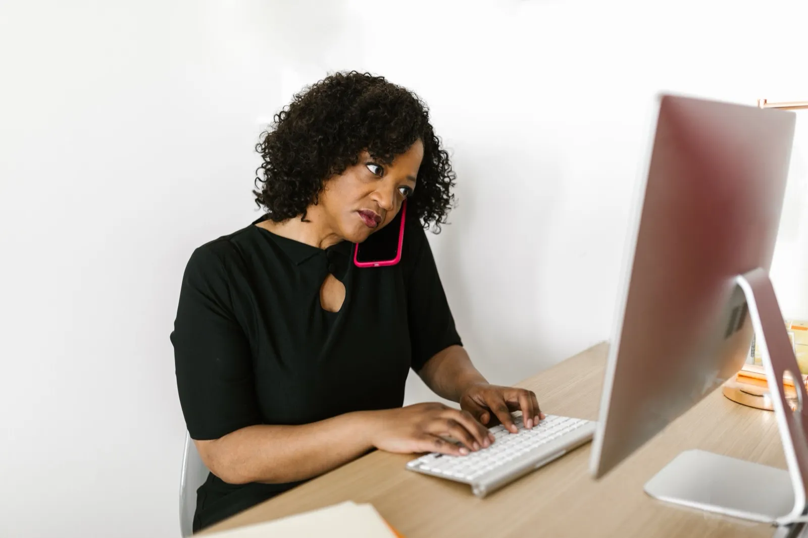 A woman on a phone call types at a desktop computer in a bright office