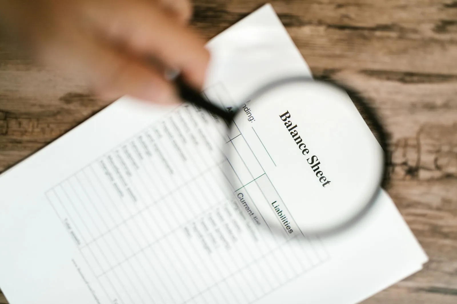 A hand holds a magnifying glass over a printed balance sheet on a wooden table