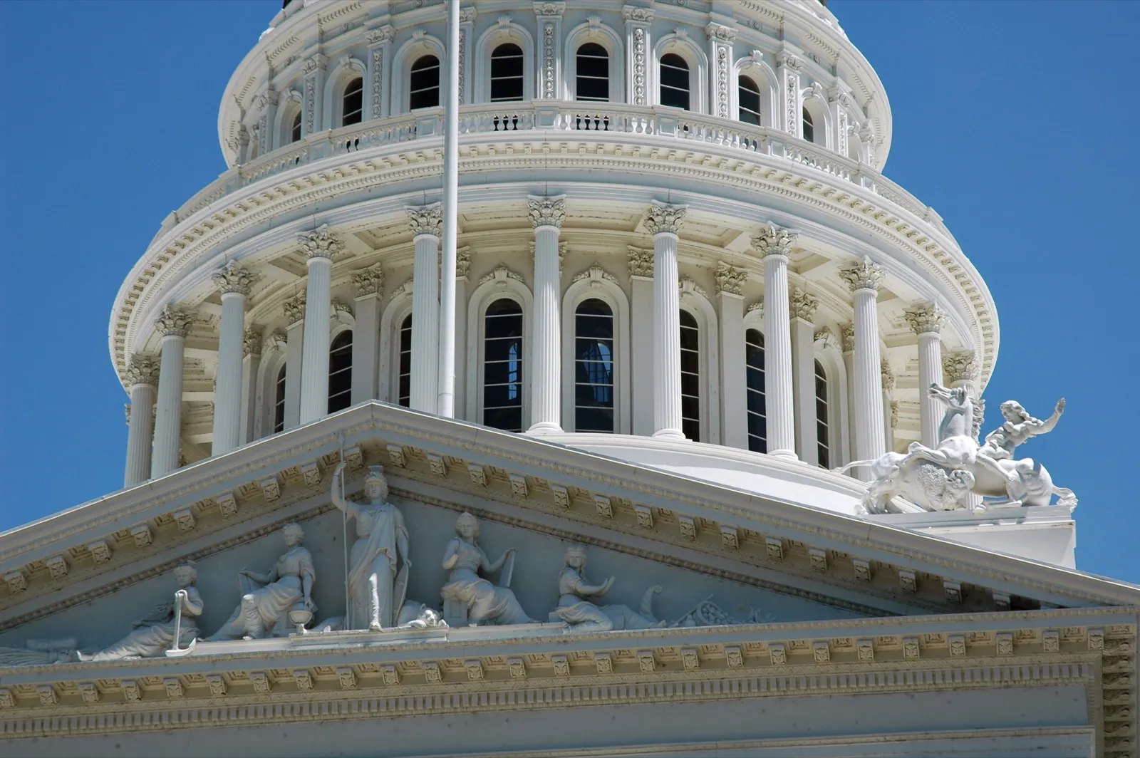 The white dome of the California State Capitol in Sacramento above its pediment of carved figures, under a clear blue sky