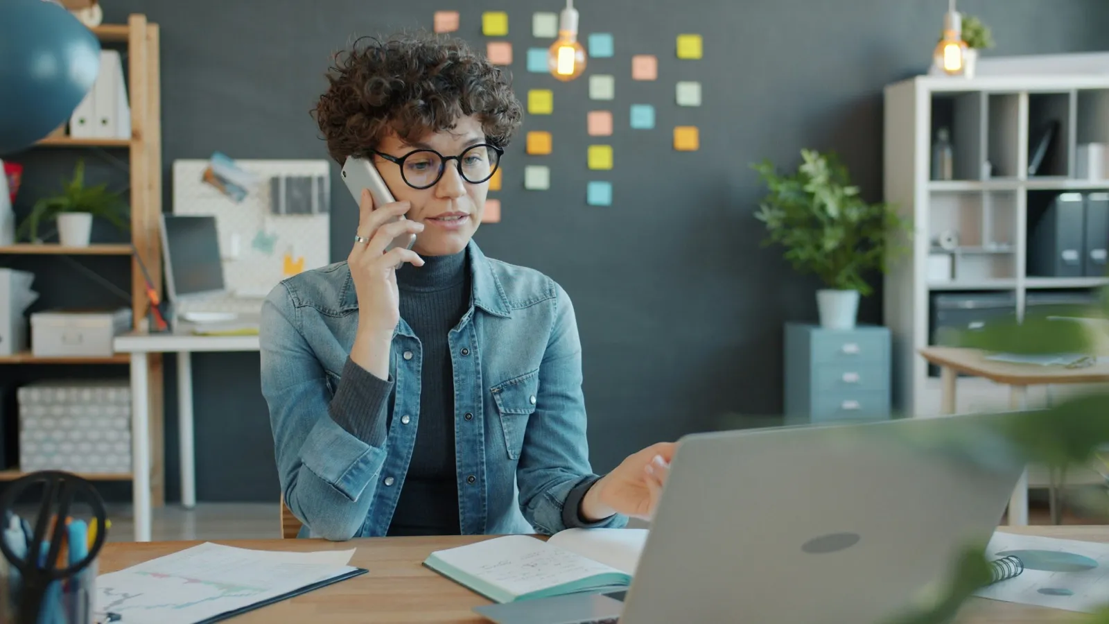 A woman in glasses talking on a phone at an office desk while reading her open laptop