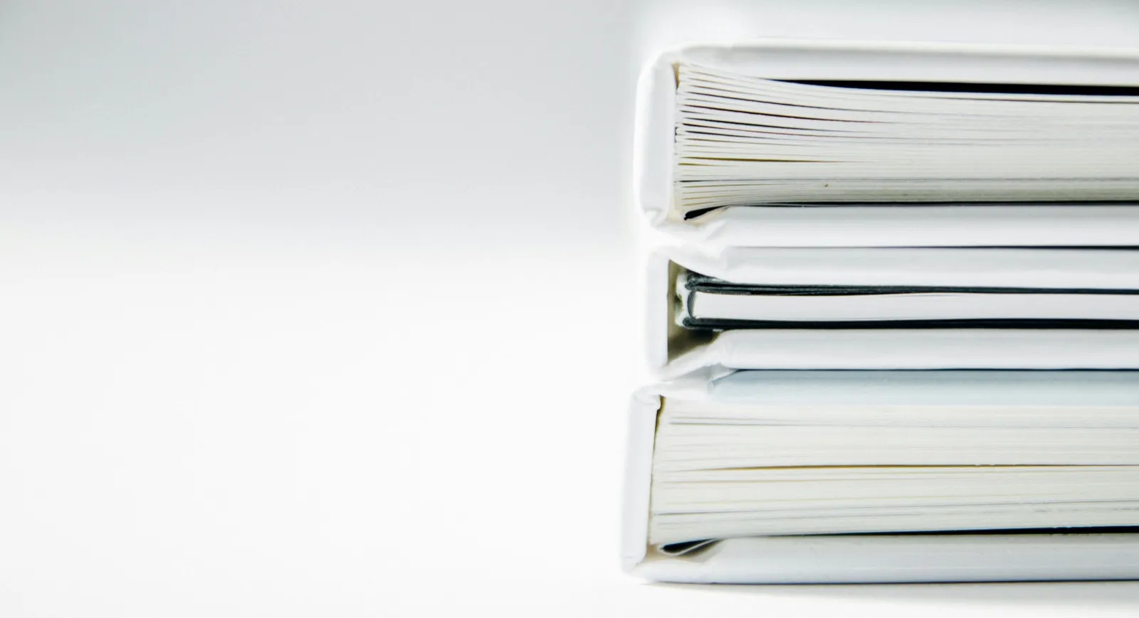 A stack of three thick white binders on a white table, seen from the side with their pages showing