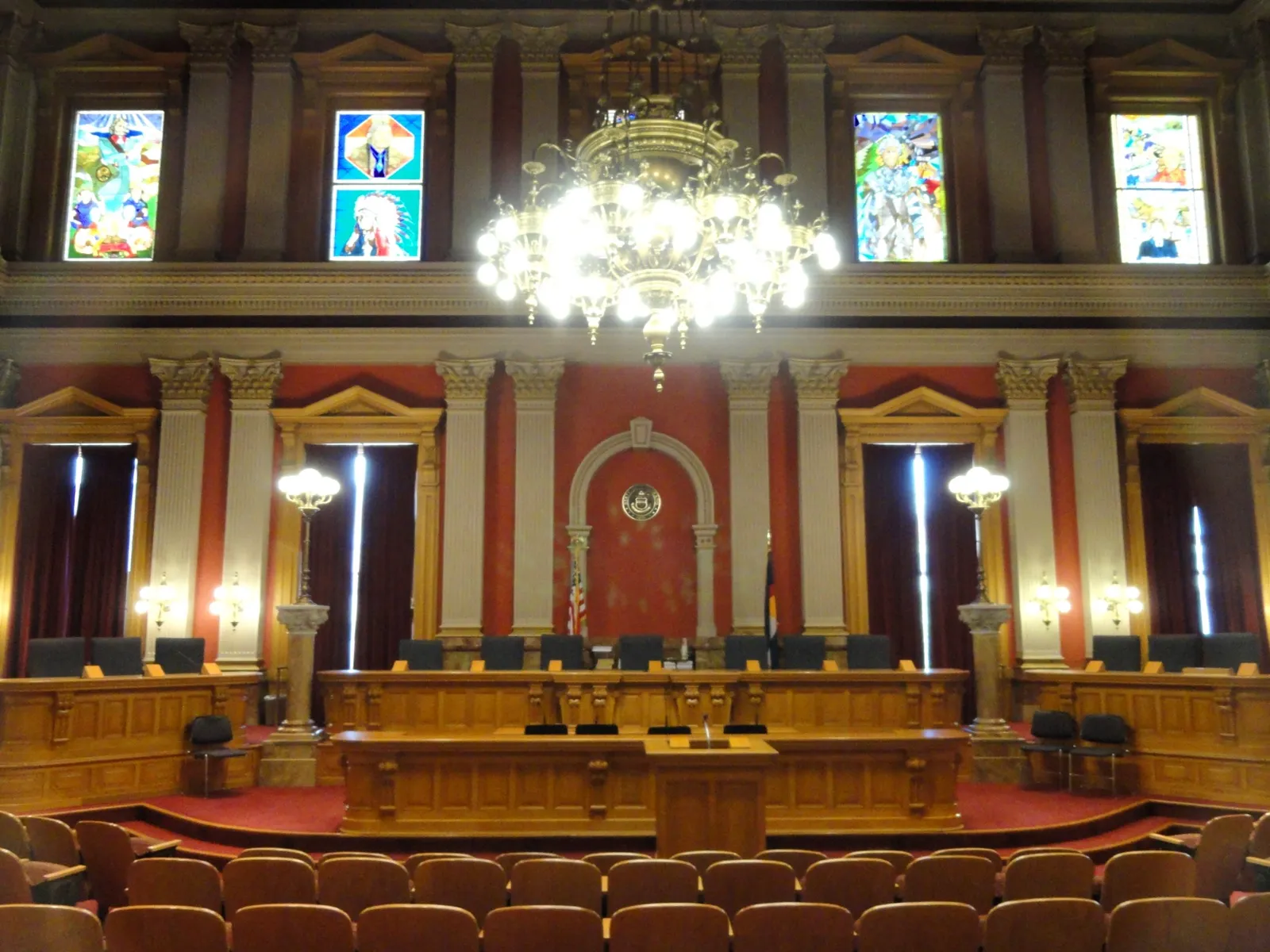 An empty hearing room in the Colorado State Capitol, with rows of seats facing a long wooden dais, red walls, a chandelier, and stained glass windows