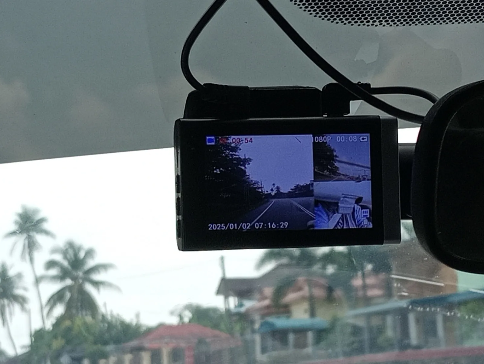 A dash camera mounted on a car windshield, its screen showing the road ahead and a smaller view of the inside of the car