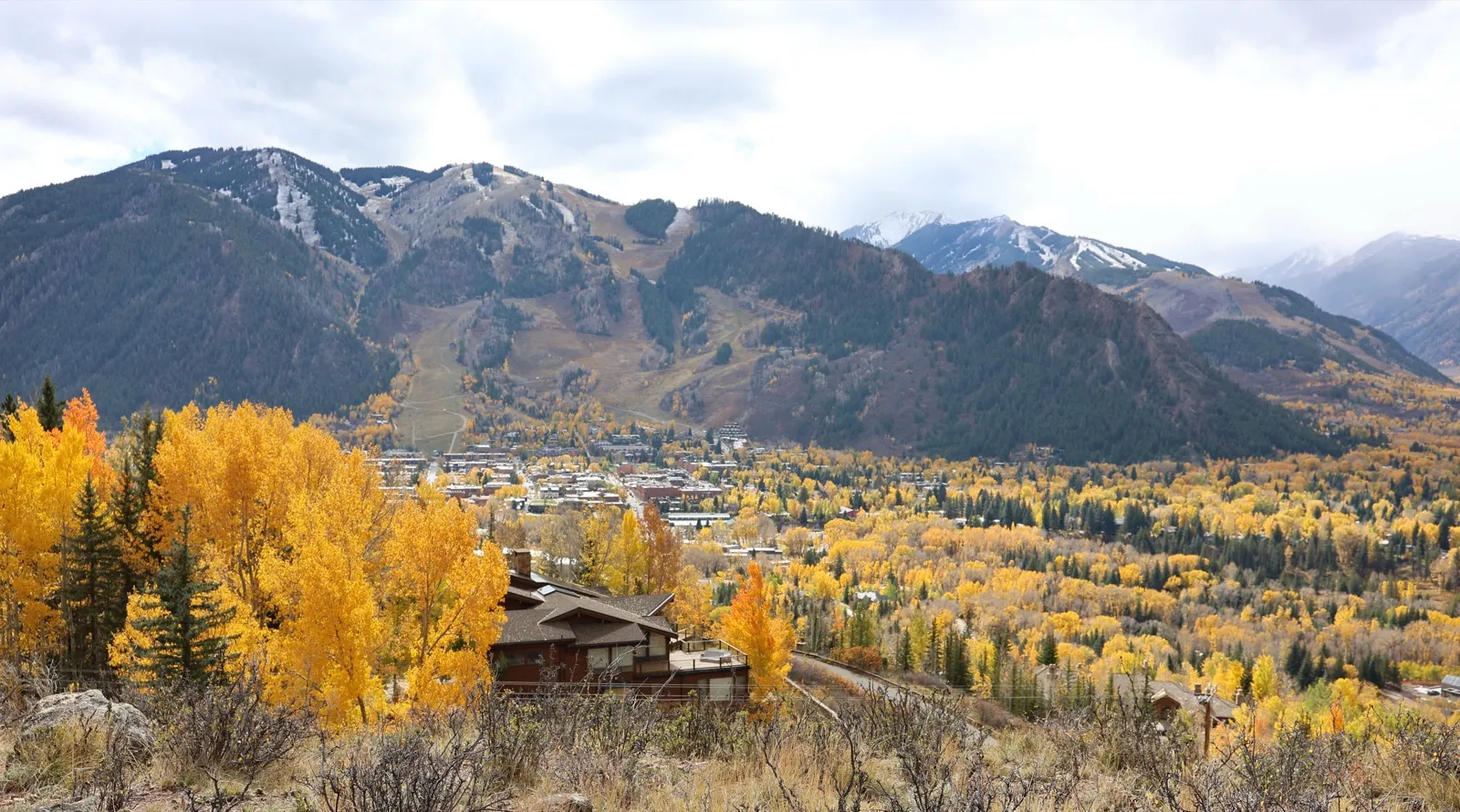 A Colorado mountain town spread across a valley of yellow aspen trees below forested ski slopes and snow-dusted peaks in autumn