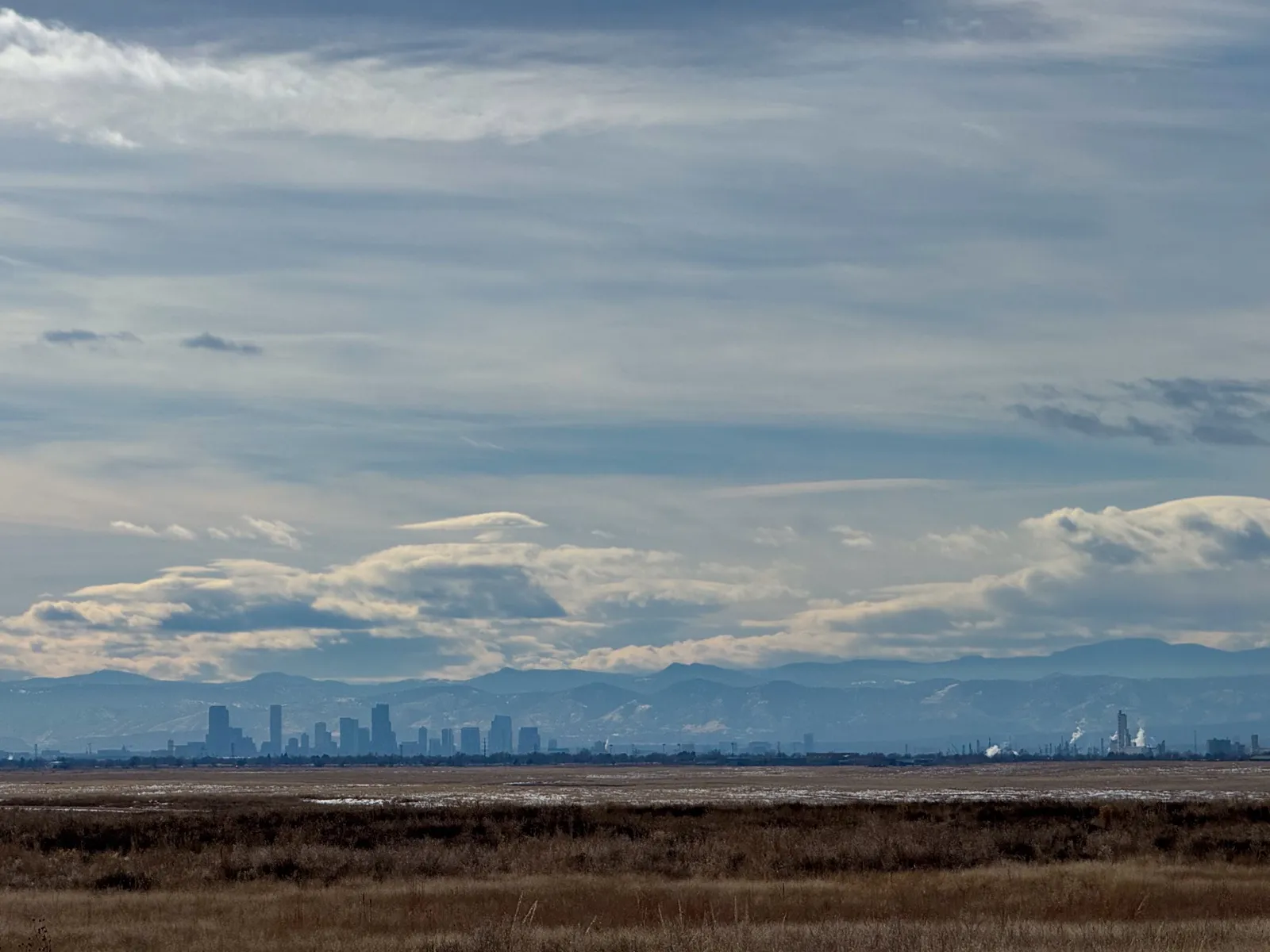 Downtown Denver's towers and the Front Range mountains seen across open prairie grass under a wide, cloudy sky