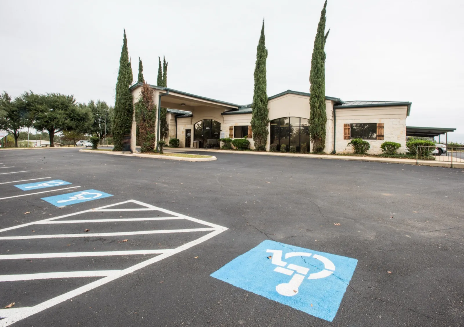 A one-story rural health center building with tall cypress trees out front and blue accessible parking spaces in the empty lot