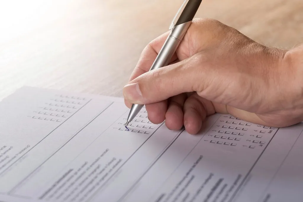 A hand holding a silver pen marks a box on a printed paper form lying on a wooden desk