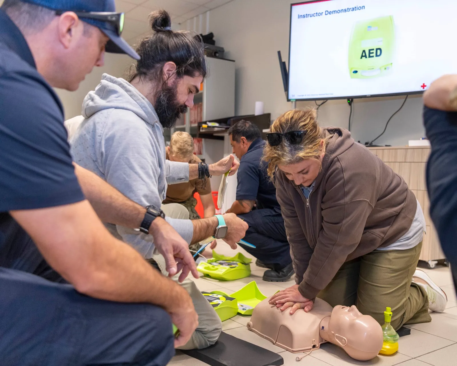 Adults kneel on a classroom floor in a CPR and AED course while a student practices chest compressions on a mannequin