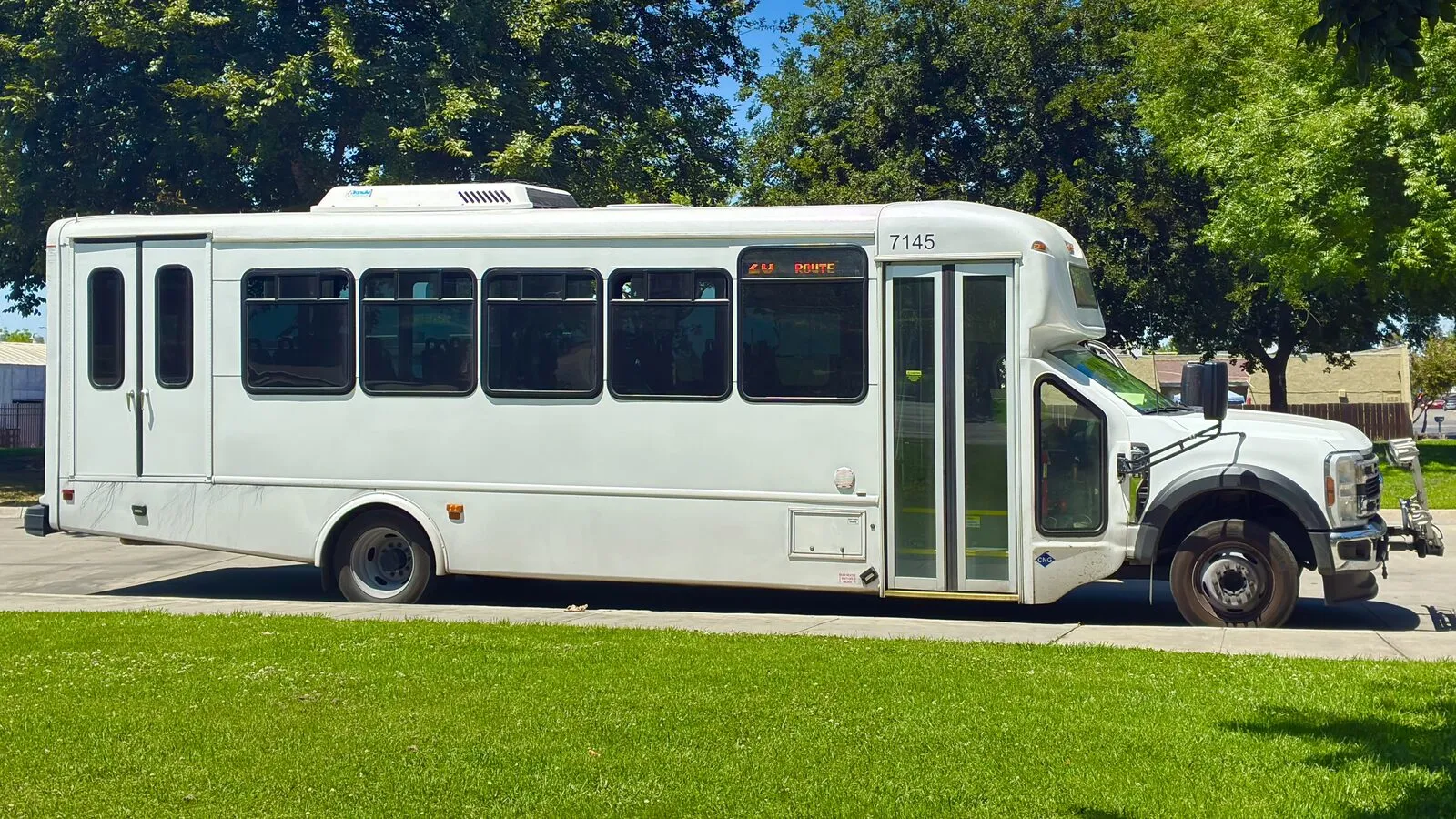 A white cutaway shuttle bus with a front passenger door, a rear double door, and a long row of side windows, parked beside a lawn