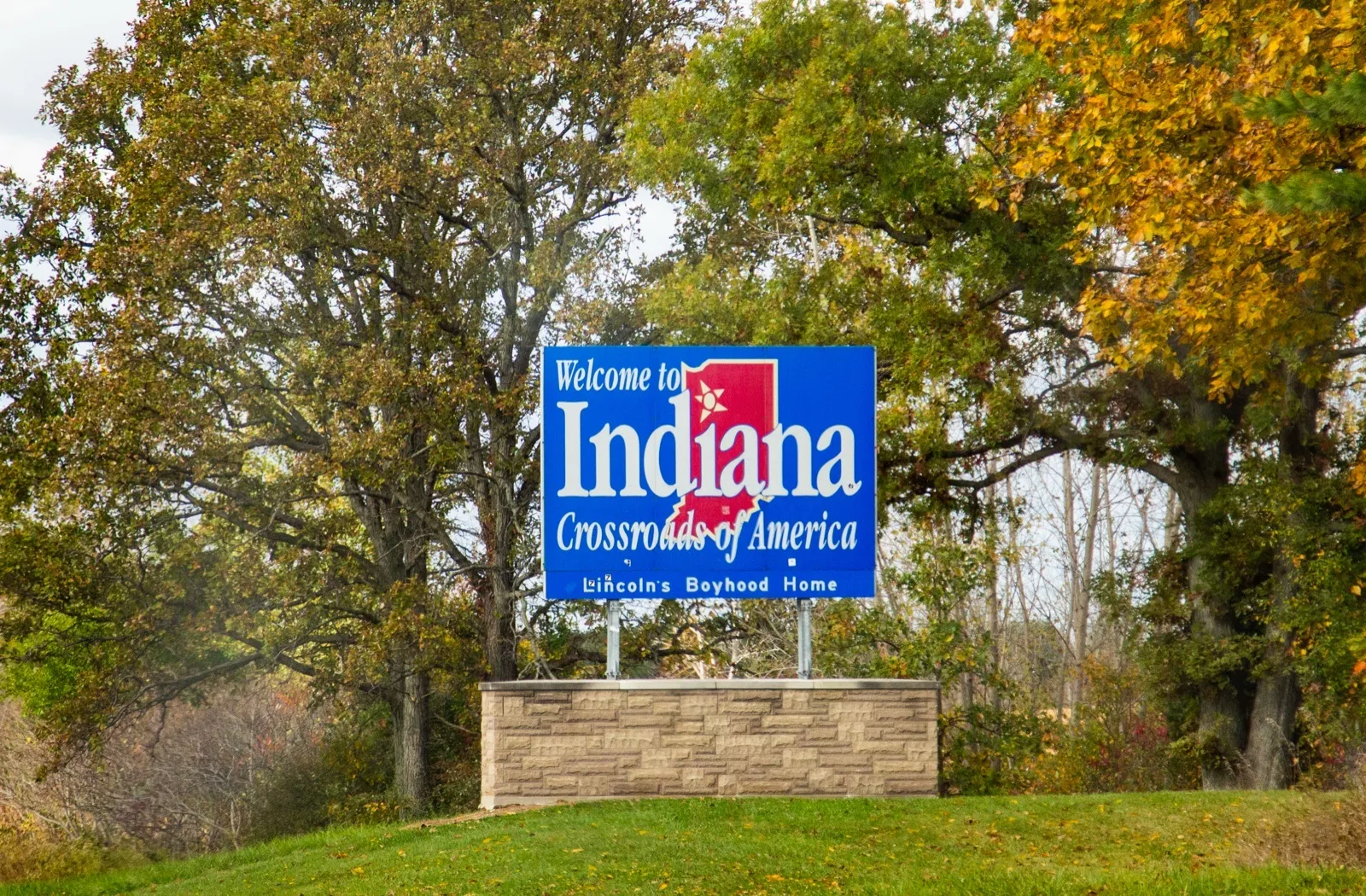 A blue Welcome to Indiana, Crossroads of America state line sign on a stone base on a grassy slope, with autumn trees behind it