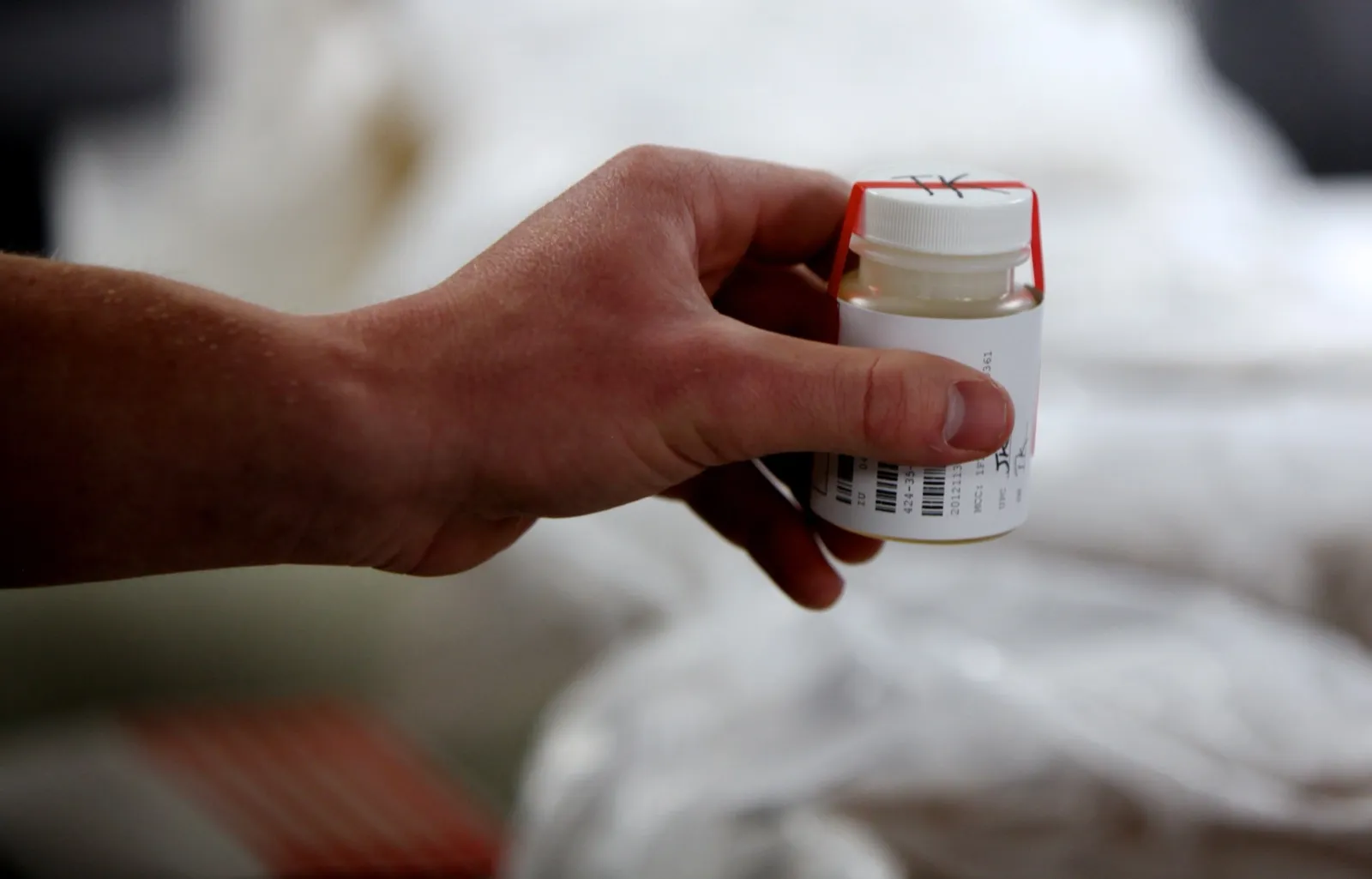 A hand holding a sealed urine specimen bottle with a red tamper seal and a bar-coded label
