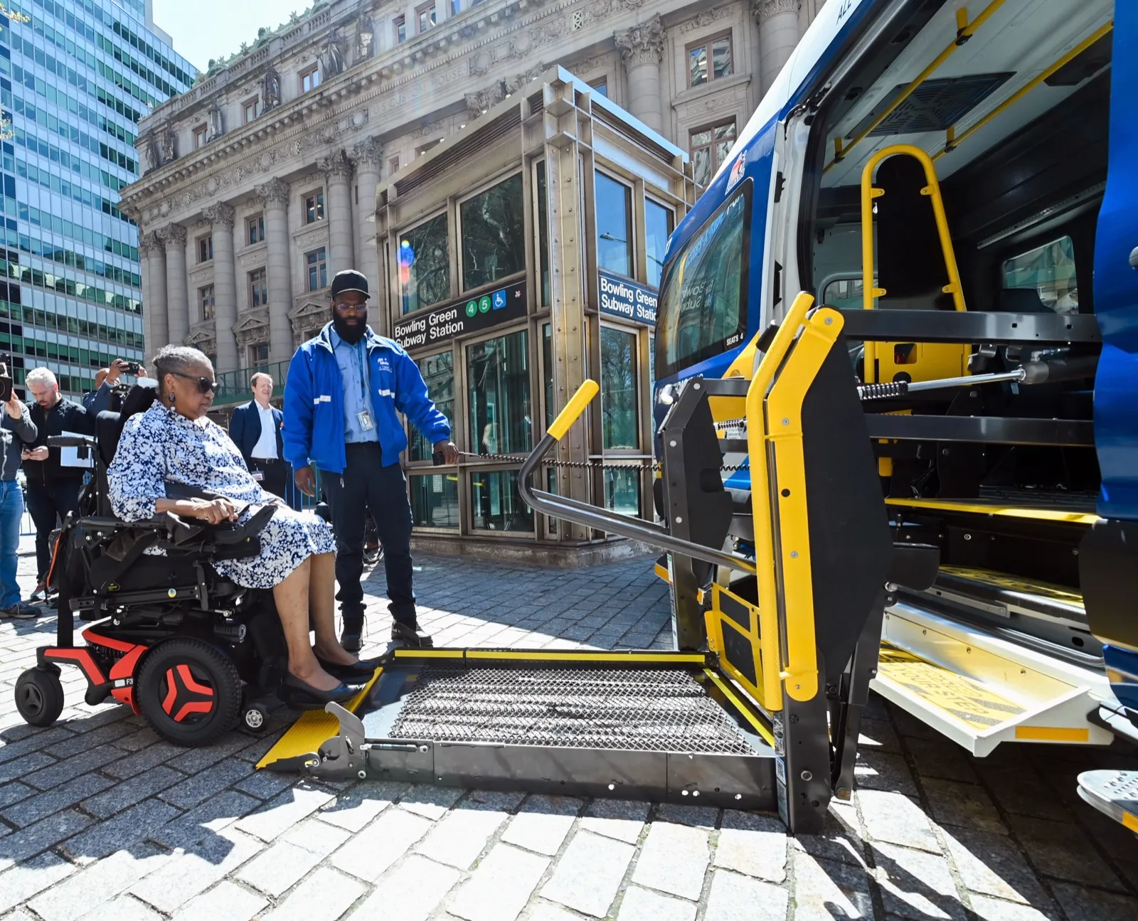 A woman in a power wheelchair waits beside the lowered side lift of an all-electric paratransit van in New York City