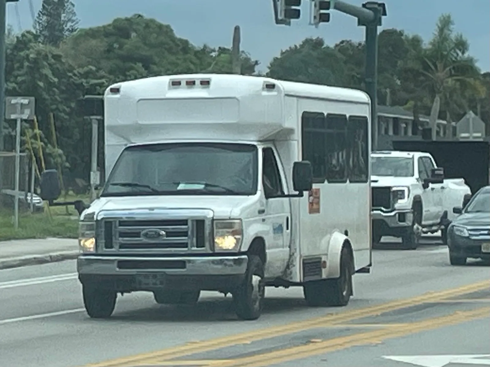 A small white cutaway bus with a passenger cabin drives along a road past palm trees in Fort Pierce, Florida