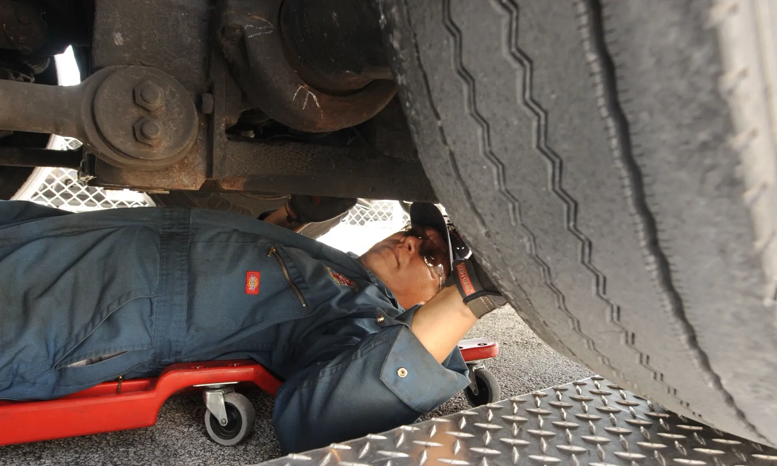 An inspector lies on a red creeper under a bus, checking the axle and suspension beside a rear tire