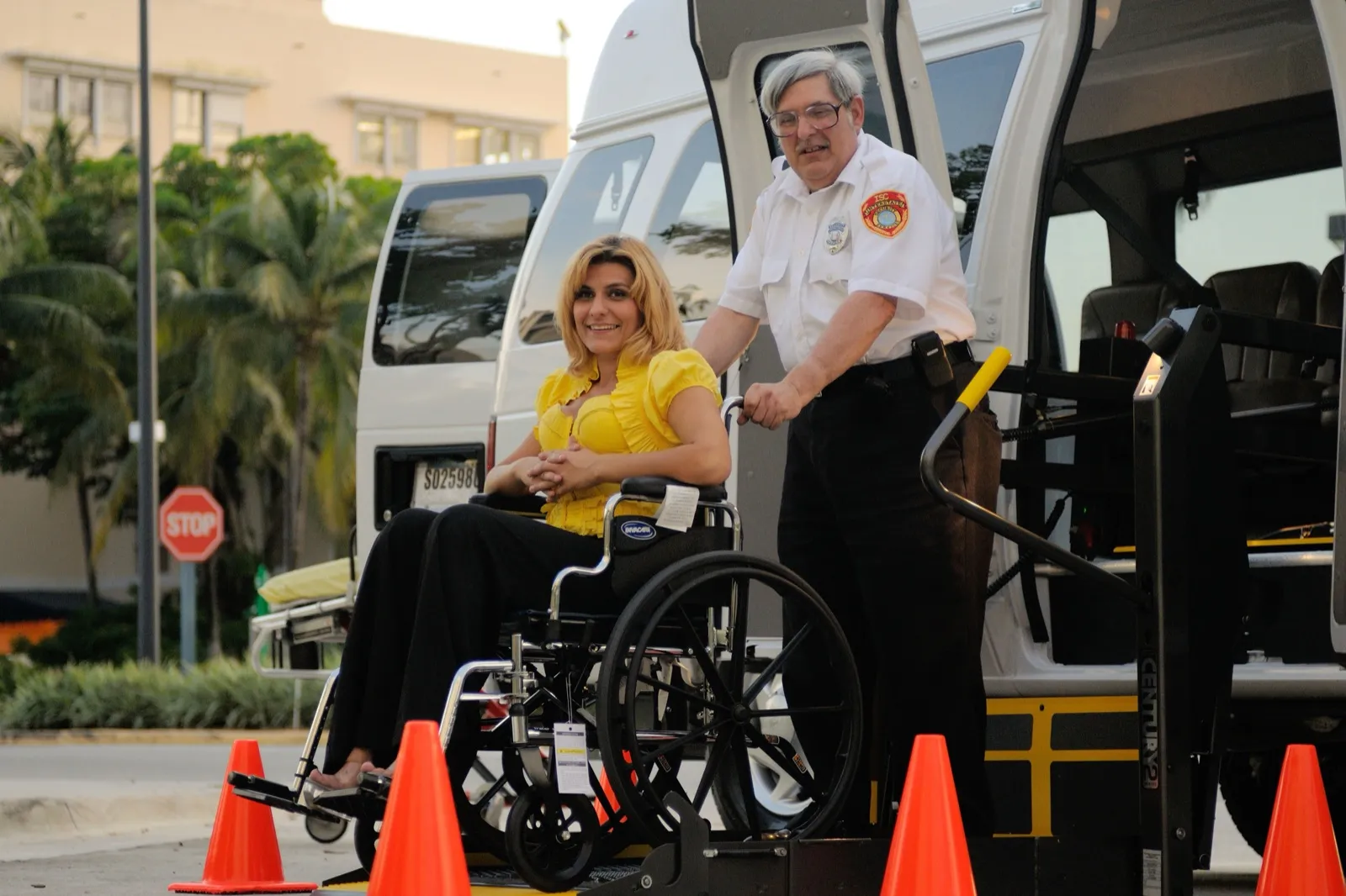 A driver standing behind a smiling woman in a wheelchair on the lift of a white accessible van