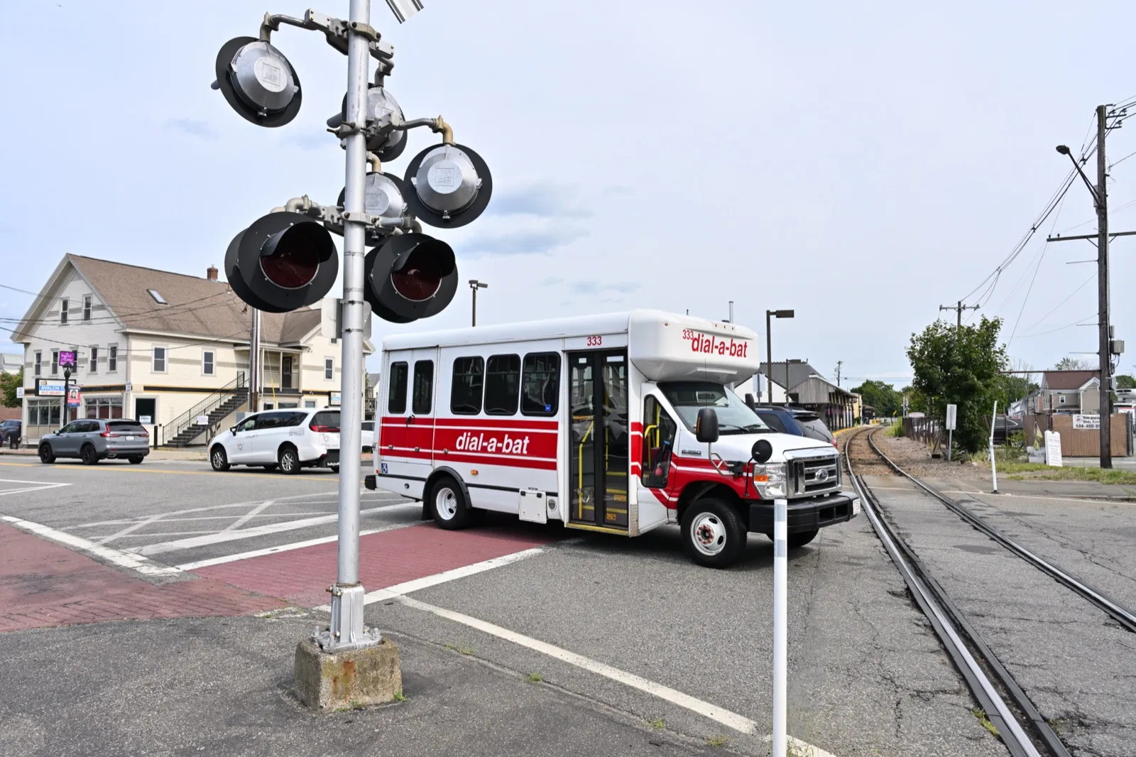 A white and red public transit dial-a-ride bus crossing a railroad track at a town intersection, with crossing signals in the foreground