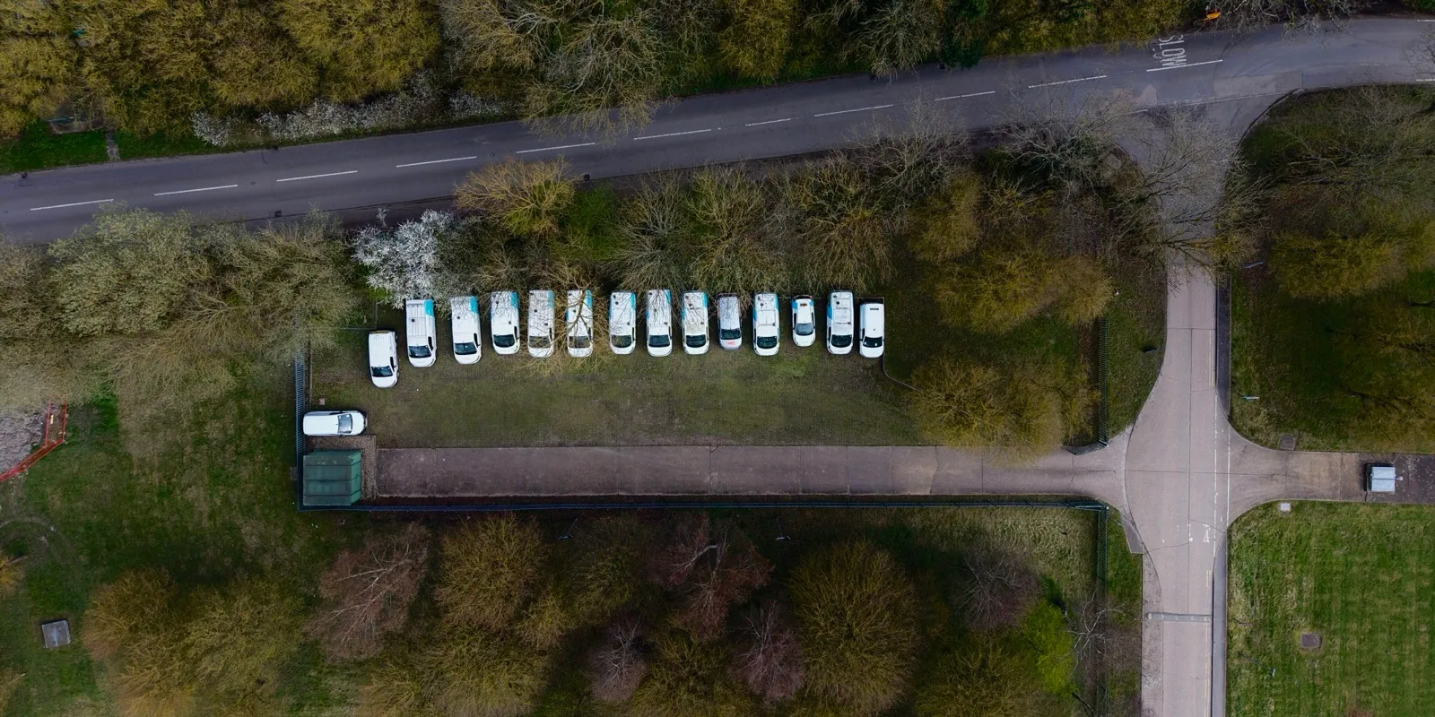Aerial view of a long row of white vans parked side by side on a grass lot beside a country road
