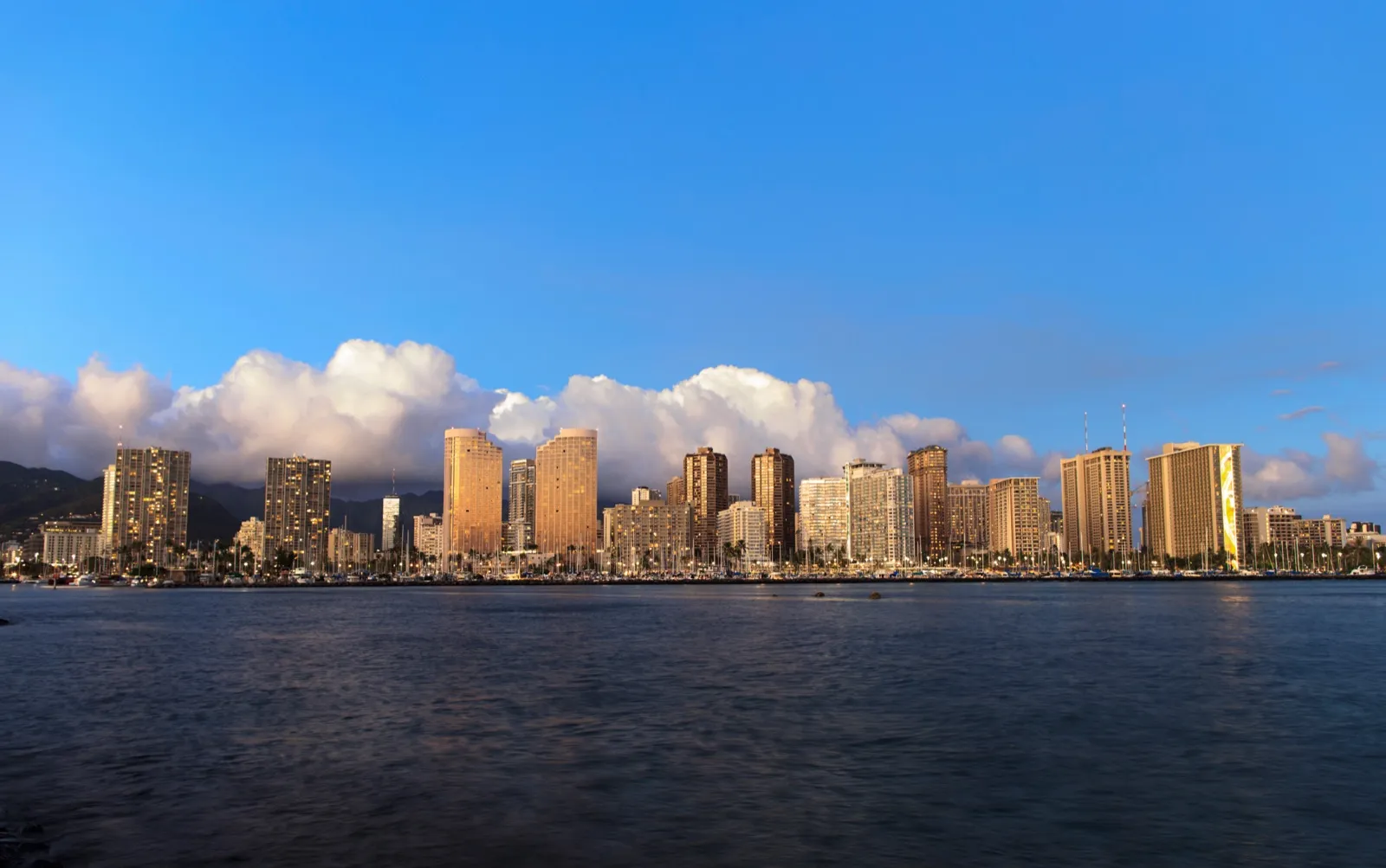 Honolulu high-rise towers and a marina along the water in late afternoon light, with mountains and white clouds behind