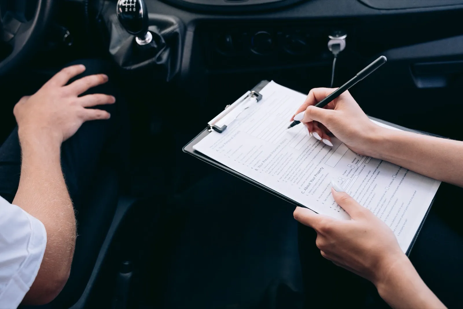 Hands filling in a paper form on a clipboard inside a vehicle, next to the gear shift