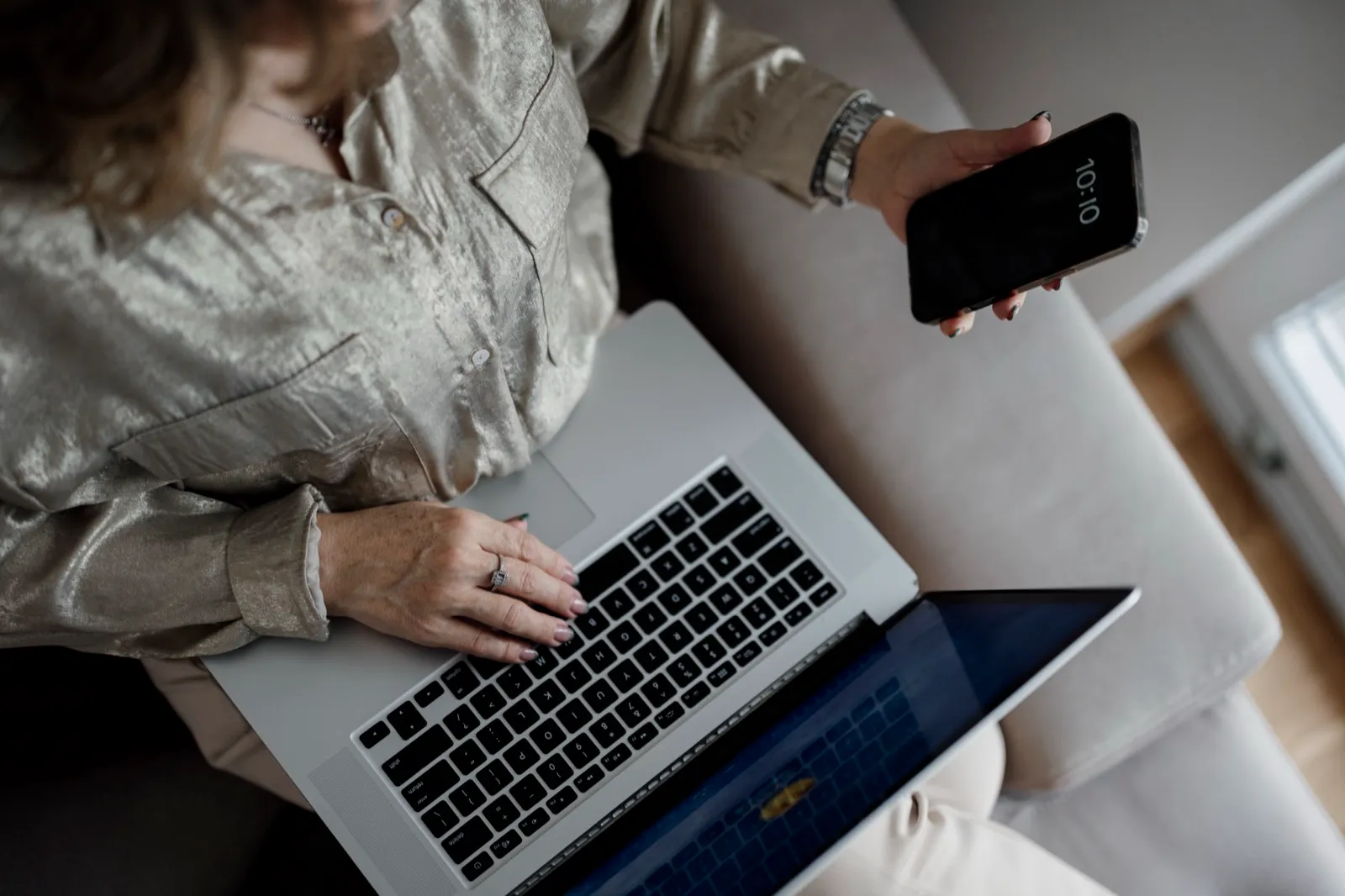 A person on a couch types on a laptop while holding a smartphone in the other hand