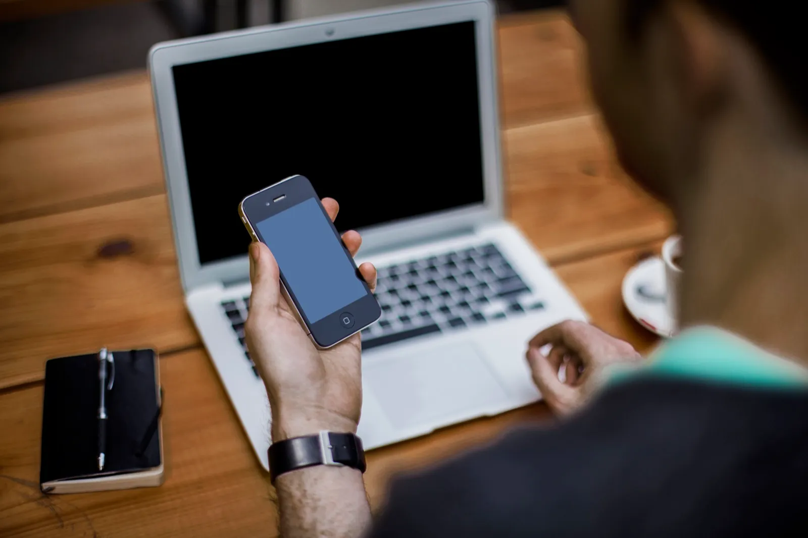 A person at a wooden desk holds a smartphone beside an open laptop, with a notebook and pen to one side