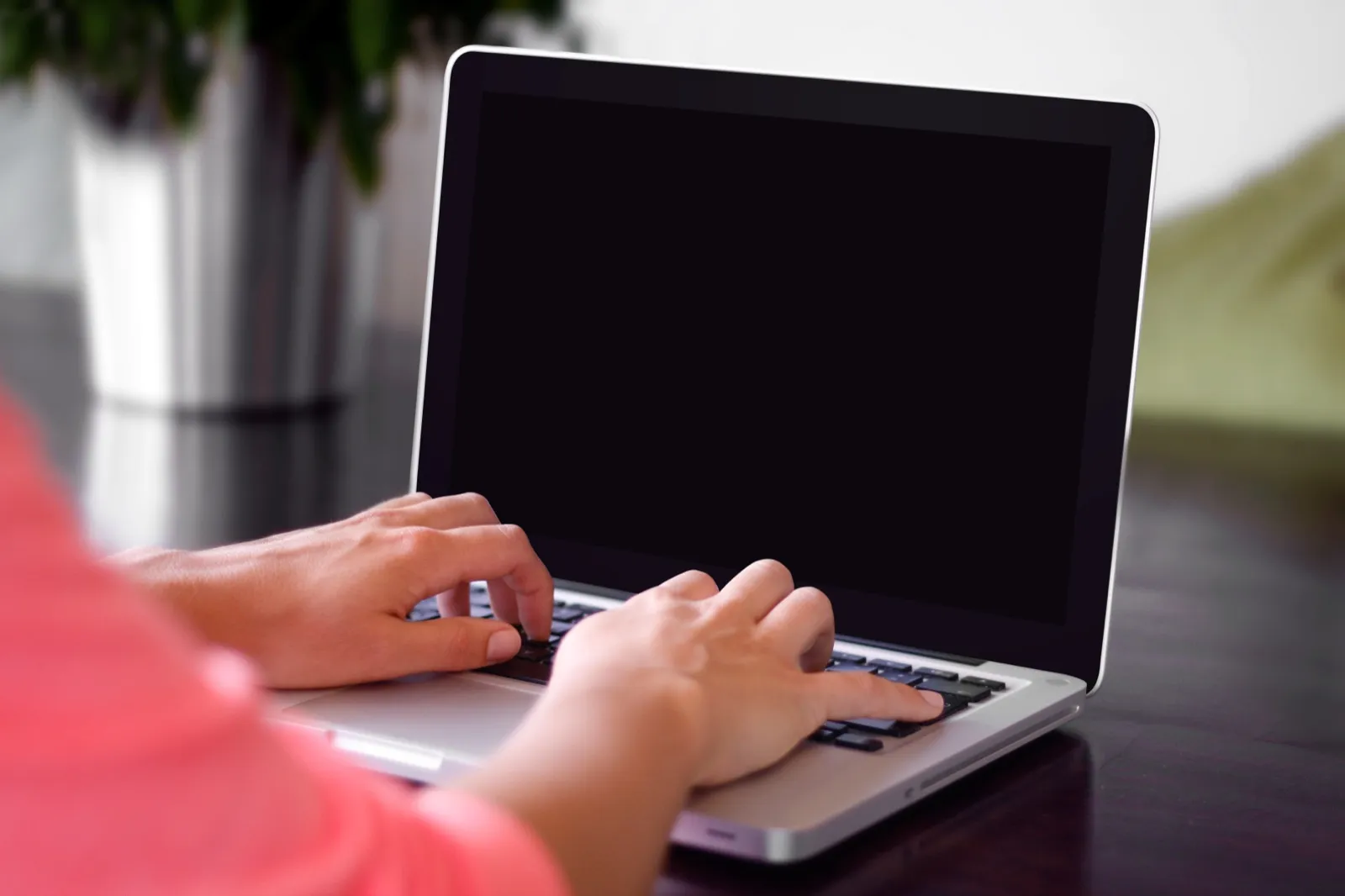Two hands typing on a silver laptop at a dark table, with a green plant blurred in the background