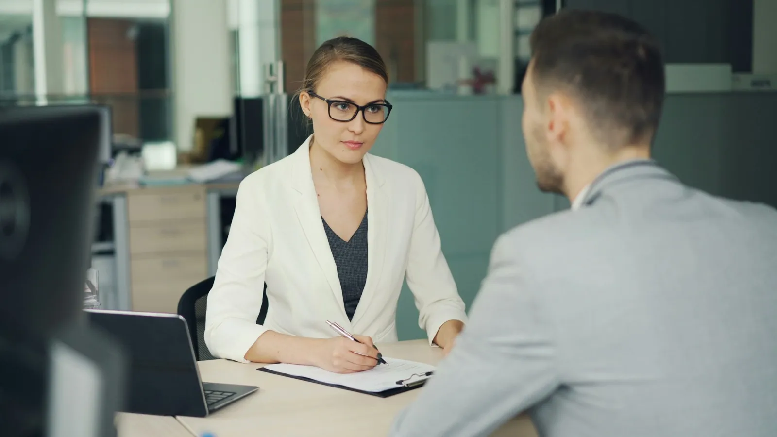 A woman in glasses takes notes on a clipboard while interviewing a job candidate across an office desk