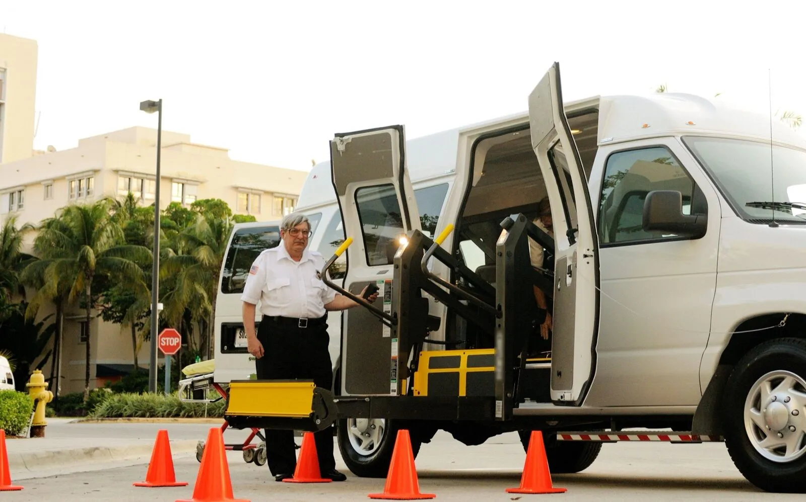 A driver in a white uniform shirt stands beside the lowered wheelchair lift of a white van, with orange cones set out around it