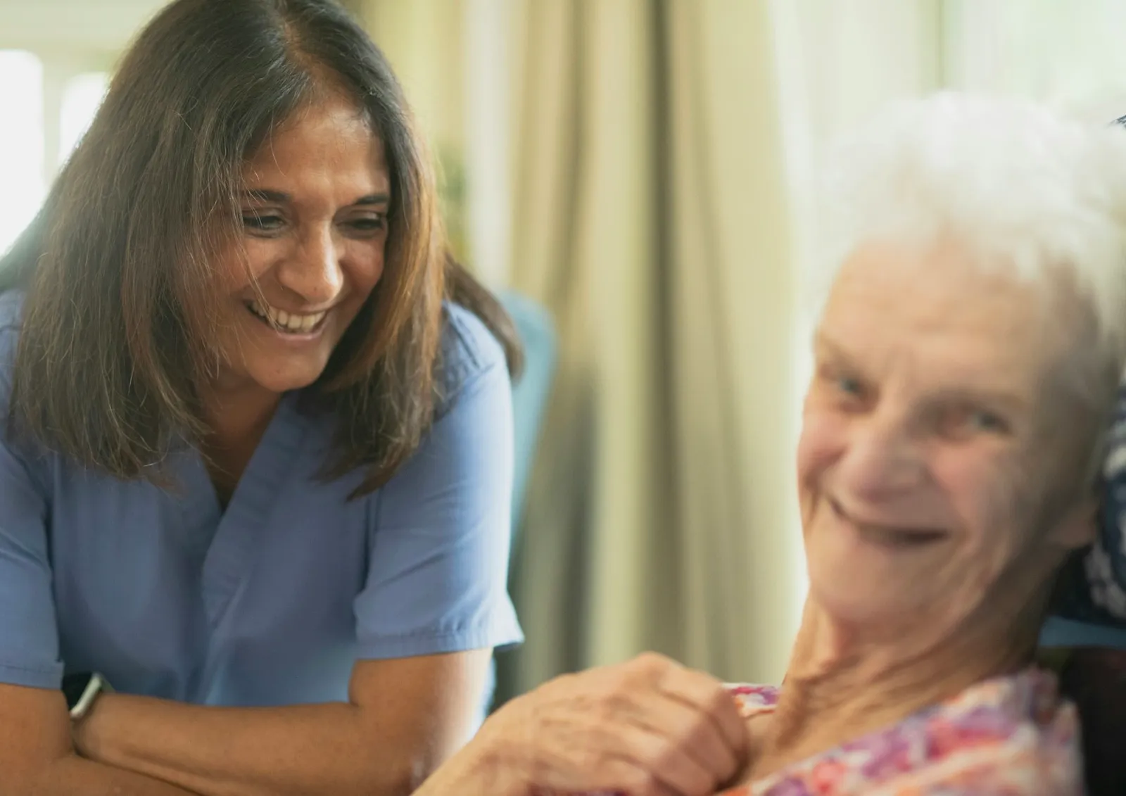 A smiling nurse in blue scrubs leans in beside an older woman with white hair who is resting in an armchair and smiling back