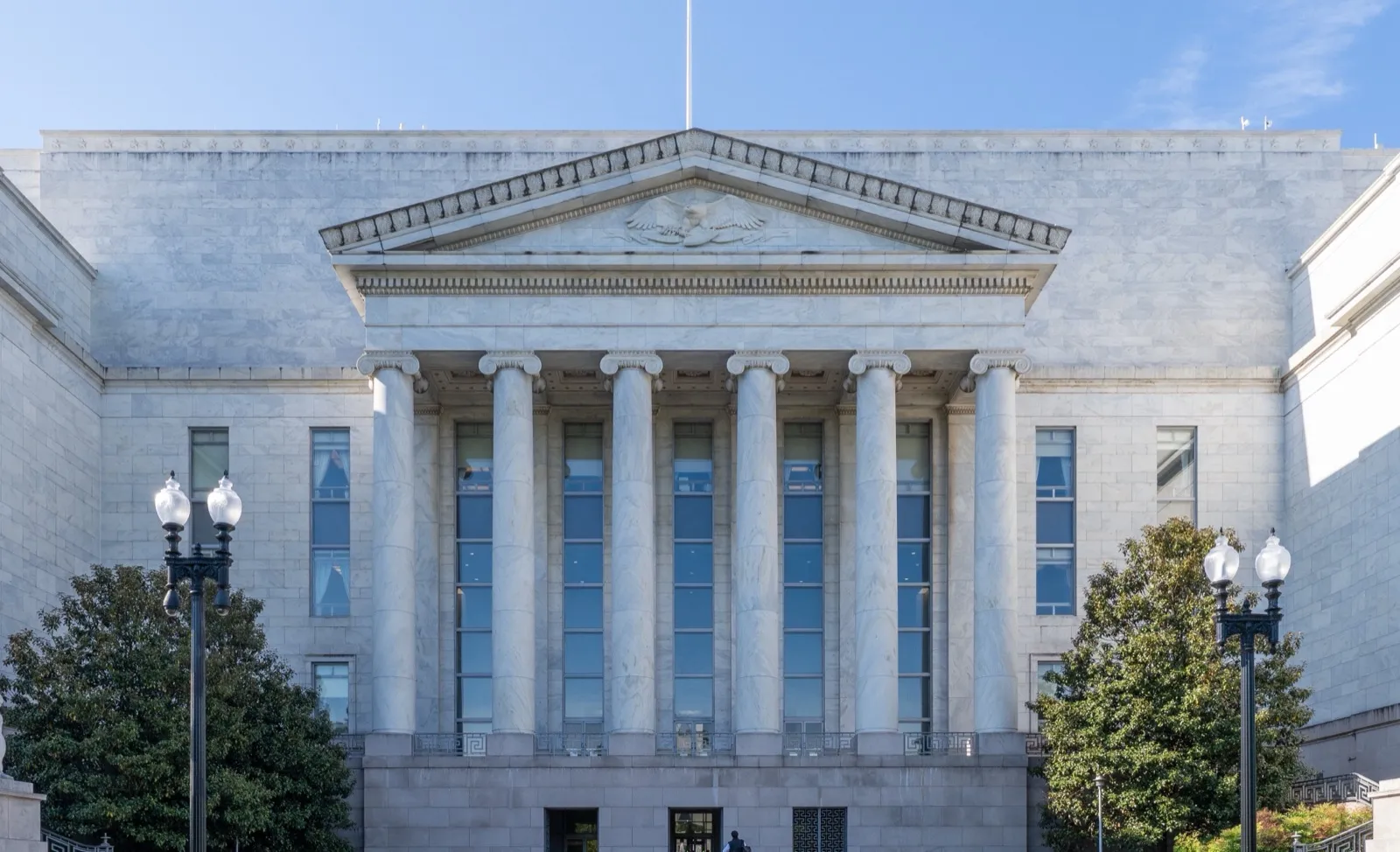 The white marble front of the Rayburn House Office Building in Washington, D.C., with six tall columns under a carved eagle, lamp posts, and trees