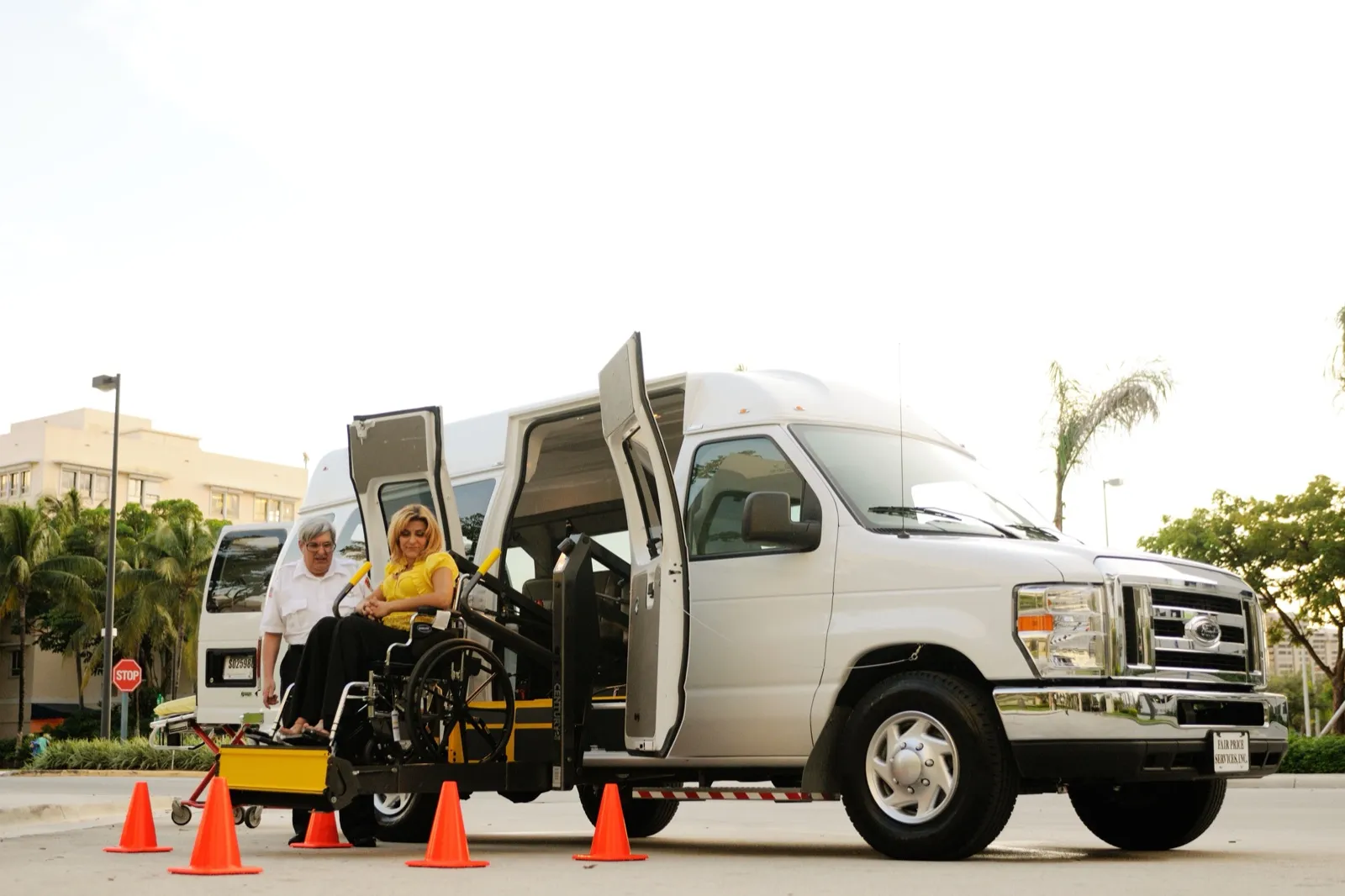 A woman in a wheelchair rides the lift of a white accessible van while a driver in a white uniform shirt stands beside her