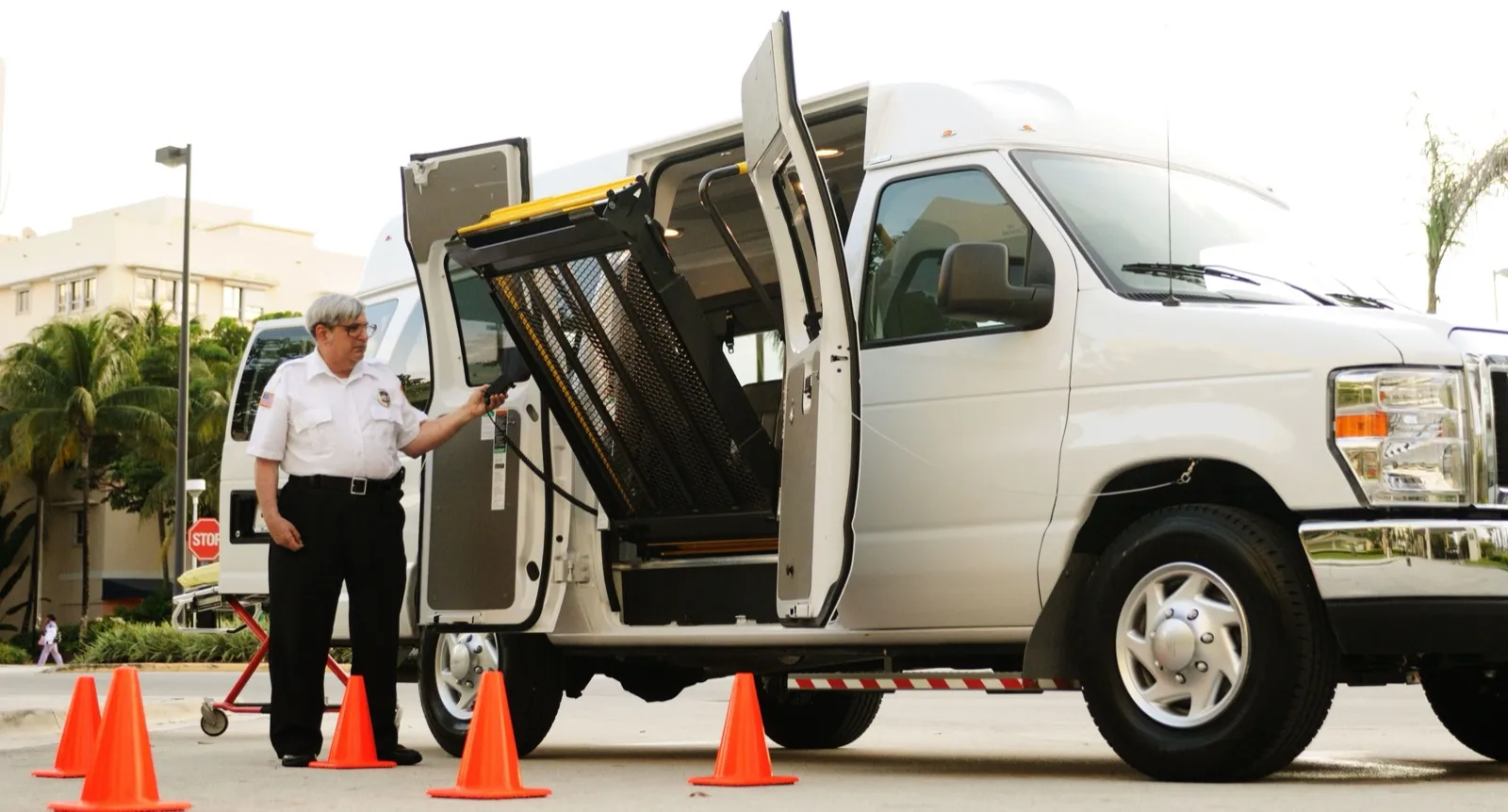 A man in a white uniform shirt works the controls of a folded wheelchair lift at the open side doors of a white high-roof van, with orange cones around it