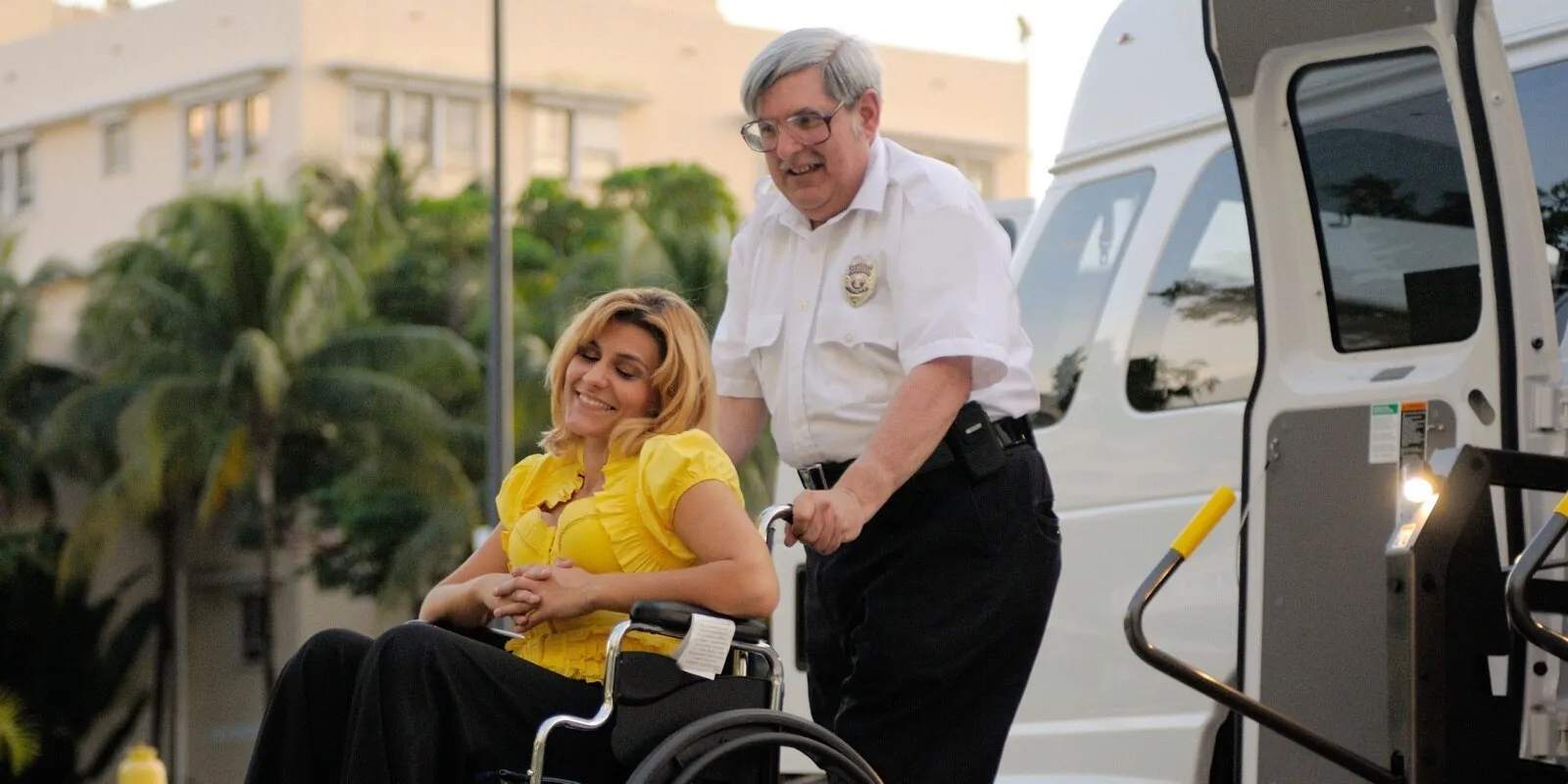 A smiling man in a white uniform shirt pushes a woman in a wheelchair toward the open lift of a white van