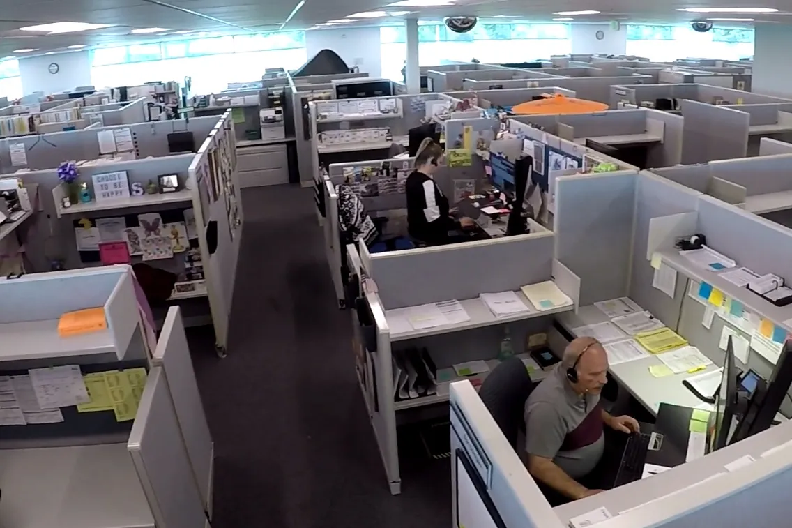 Call center staff wearing headsets work at computers in rows of cubicles in a large open office