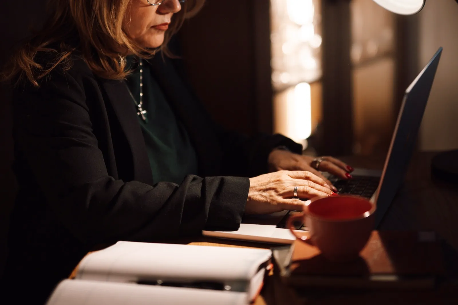 A woman typing on a laptop at a desk in a dim home office, with open notebooks and a red coffee cup beside her